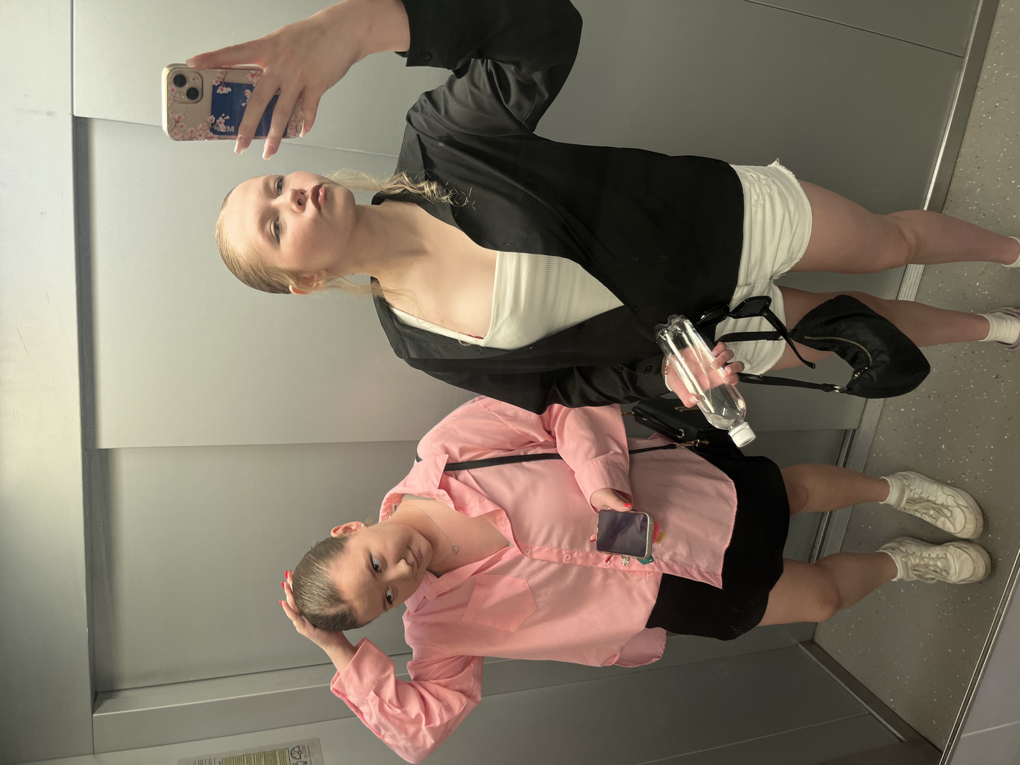 Two young women are taking a mirror selfie in an elevator. The woman on the left is wearing a pink button-down shirt and a black skirt. She has her hand on her head and is holding a smartphone in her other hand. The woman on the right is wearing a white tank top, a black open shirt, and white shorts. She is holding a bottle of water and a smartphone in her hands, and is making a pouty face towards the camera. The elevator appears to be in a modern building. The location is not identifiable as Perm, Russia.