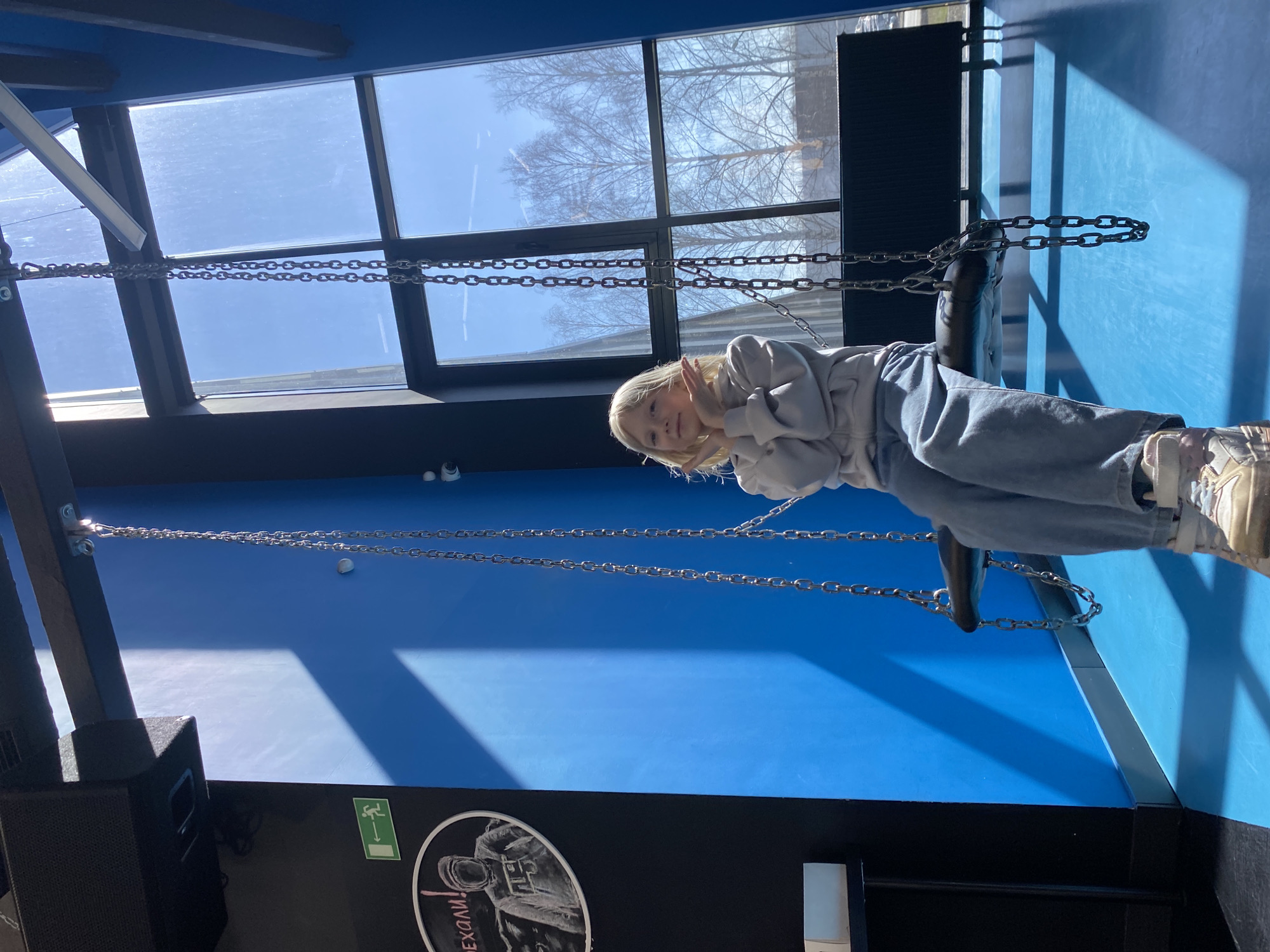 A full shot of a young blonde-haired girl in casual clothing sitting in a swing suspended by chains from a ceiling, likely indoors given the presence of a large window with visible bare trees outside. The room features bright blue walls. Below, on a black section, is a circular logo with the text "Ехали!" and a stylized drawing. This scene is likely to be in Izhevsk, Russia.