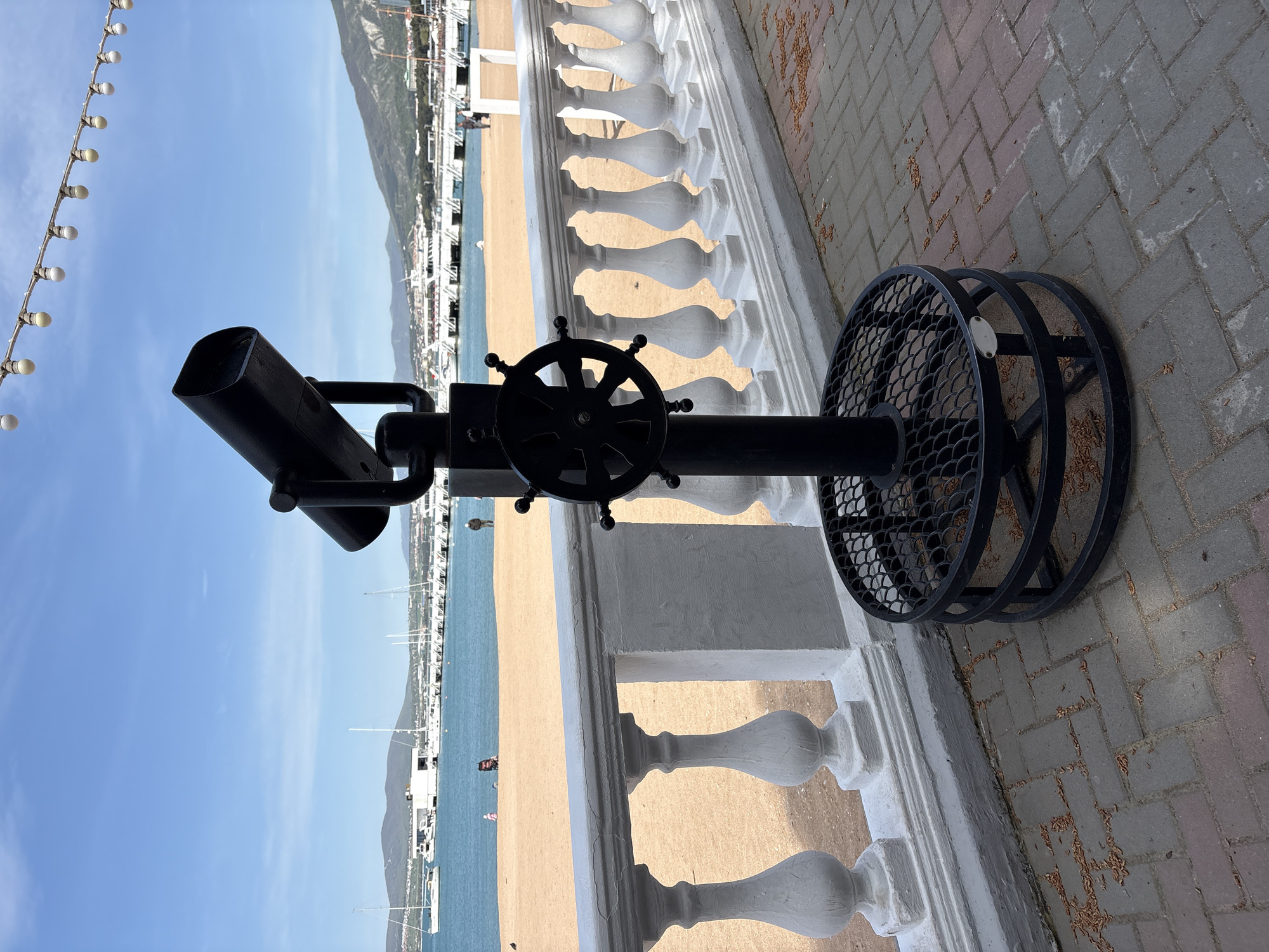 This image captures a vibrant seaside promenade in Gelendzhik, Russia, on a bright, clear day. In the foreground, a black metal coin-operated telescope or binocular viewer stands prominently, featuring a ship's wheel-like handle for adjustment. Its circular, grated base rests on a paved walkway composed of interlocking grey and reddish-brown pavers, with scattered small leaves or seeds.
A white, ornate balustrade forms a distinct horizontal line across the midground, separating the paved area from a wide, light-brown sandy beach. Beyond the beach, tranquil blue waters of a bay or harbor are visible, dotted with numerous white yachts and boats moored along a distant pier or coastline. In the far background, a majestic mountain range rises, partially obscured by coastal buildings, under a vast, cloudless blue sky.
A string of decorative white spherical lights hangs diagonally across the upper left portion of the sky, adding a festive touch. The overall impression is one of peaceful, sunny weather, likely in the morning or early afternoon. A few very distant, indistinct human figures can be seen on the beach and near the water, but no specific activities are discernible. There is no visible text in the image.