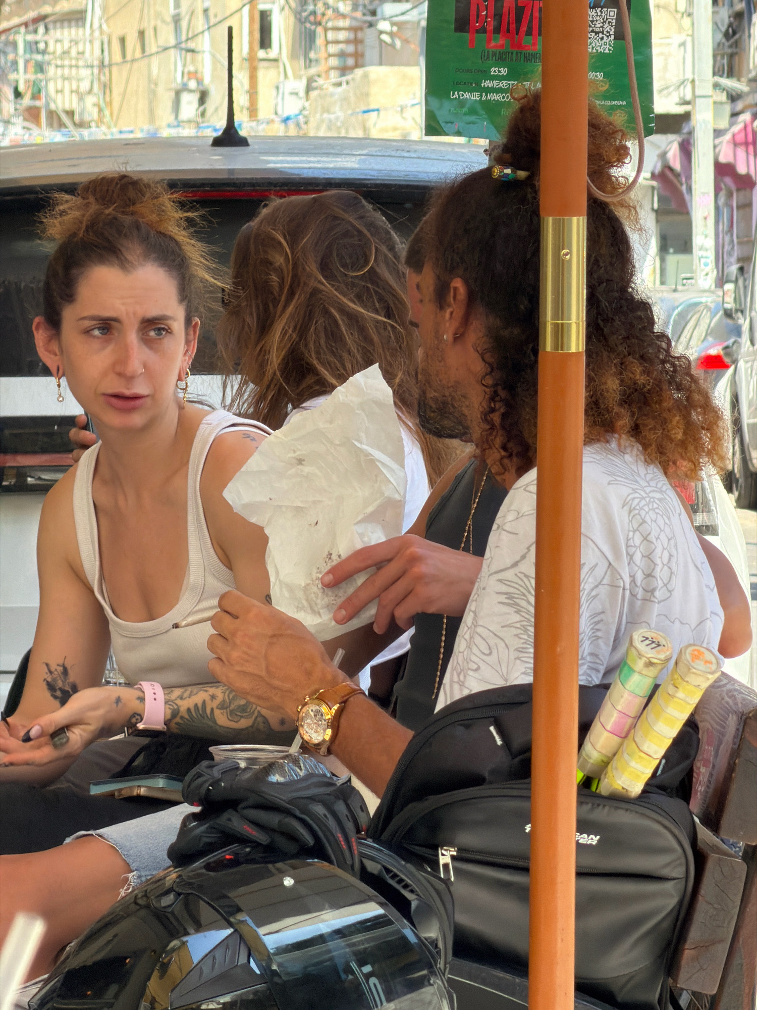 Three people are seated outdoors, possibly at a cafe or restaurant in Tel Aviv, Israel, indicated by a sign in the background that reads "PLAZA (LA PLACITA AT HAMERETZ)". The woman on the left has her hair in a bun and is wearing a white tank top. She has tattoos on her arm and is wearing a pink watch. She is looking to her left and holding a white paper object. A man with curly hair and a beard is seated to her right, wearing a dark shirt. He has a gold necklace. A third person, whose face is obscured, is seated between them. In the foreground, a black backpack with tennis rackets in it is visible. There is also a black helmet and a watch on a man's wrist. The scene appears to be in a city with buildings and parked cars in the background.