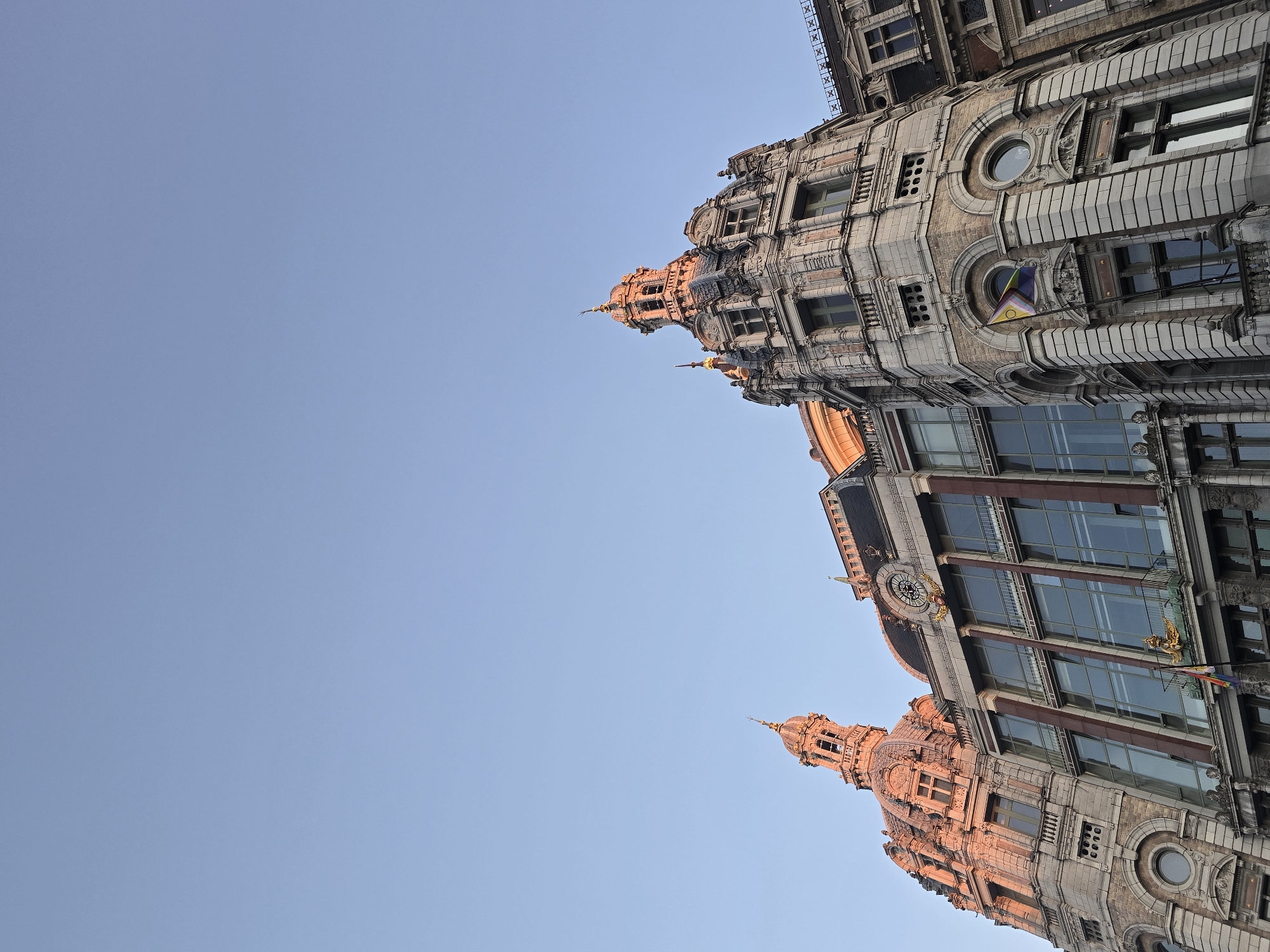 The image shows the exterior of the Antwerp Central Station in Antwerp, Belgium, captured from a low angle looking up at the grand architecture. The sky above is a clear, pale blue, suggesting either early morning or late afternoon light. The building features ornate, historical architecture with domes, towers, and intricate stone carvings. Sunlight is casting a warm glow on the upper sections of the building, highlighting the details of the facade. There are large windows and balconies on the different levels of the structure. A flag, possibly a pride flag, is visible hanging from a balcony. The scene is devoid of any visible people.