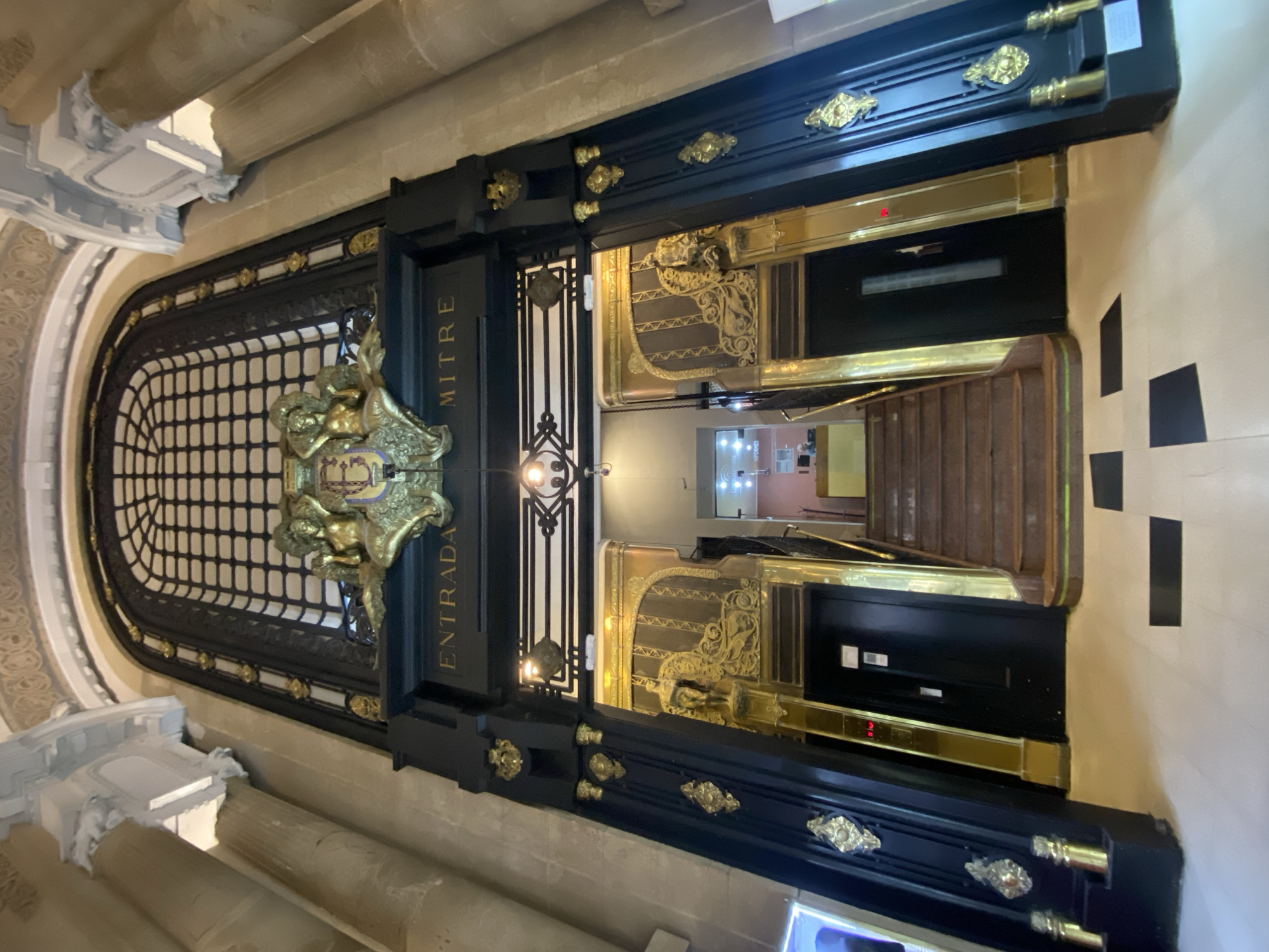 The image shows the entrance of the Palacio Mitre in Buenos Aires, Argentina. The entrance features ornate black and gold trim, with the words "MITRE" and "ENTRADA" (entrance) inscribed on either side. A decorative golden shield adorns the top of the doorway. Beyond the entrance, stairs leading to an upper level can be seen, as well as an elevator. The walls are painted in a checkerboard pattern of white and black squares. The interior space is visible beyond the entrance.