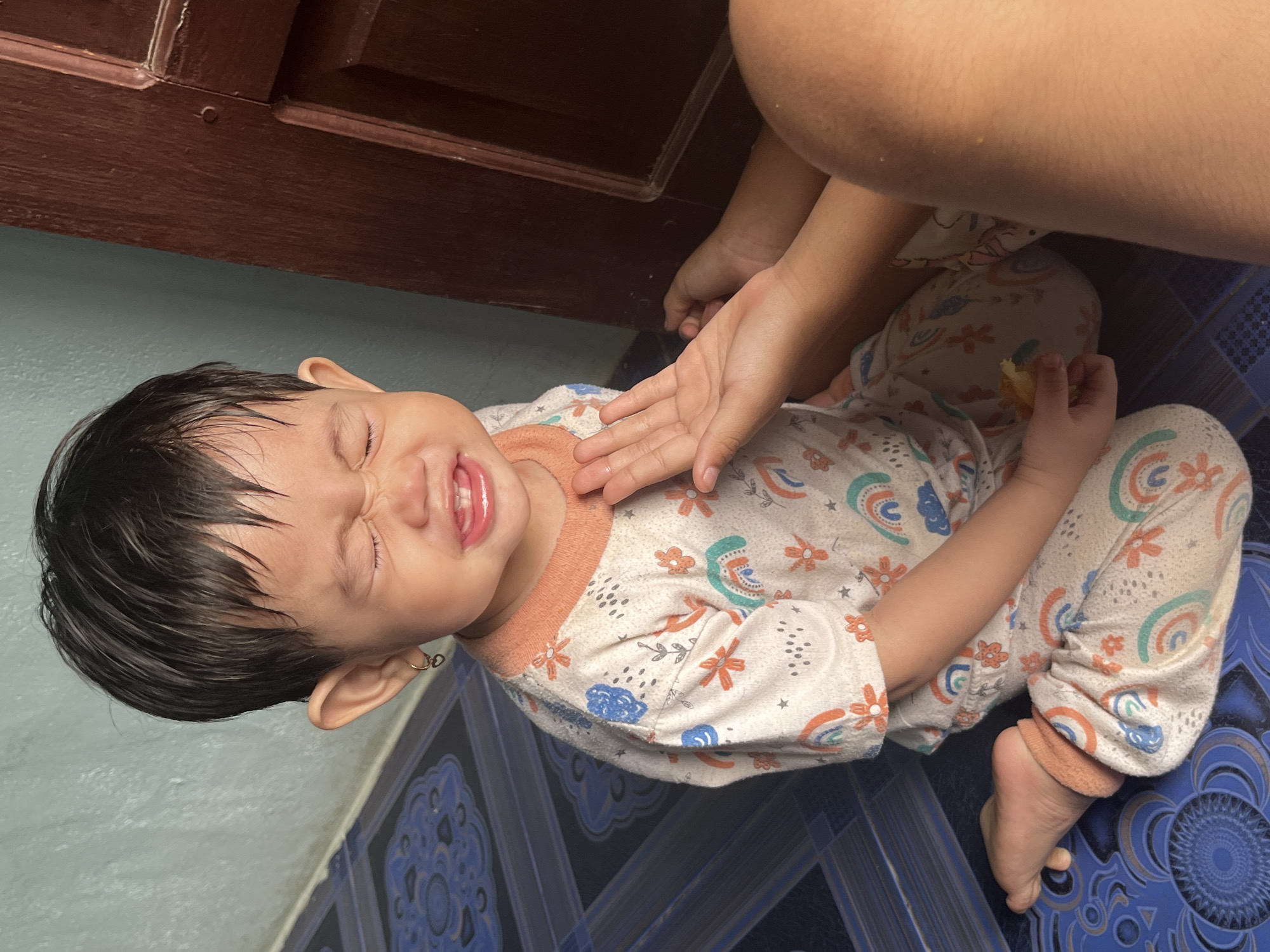 A child is crying on a patterned floor next to a wooden door. The child is wearing a white pajama set with a pattern of rainbows and flowers. The child has dark hair and is wearing a small hoop earring in their left ear. A person's hand is reaching out towards the child. The background is a light blue wall and a brown wooden door. The scene is indoors, likely in a home. The floor tiles have a blue and white pattern. The child is sitting cross-legged on the floor. There appears to be a small piece of food in the child's hand. The image is taken from a slightly elevated angle. The location is Meliau Hulu, Indonesia.
