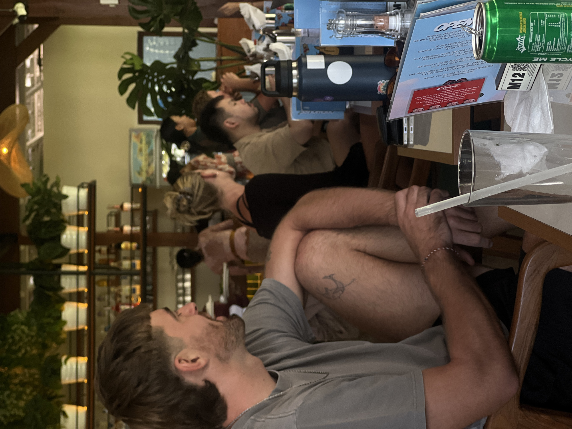 A medium shot captures a man with a mullet and beard in the foreground, seated in a restaurant in Mengwi, Indonesia. He is wearing a grey t-shirt and black shorts, and his left arm rests on his bent knee, revealing a tattoo on his thigh. His head is turned to the left, and he appears to be looking at someone outside the frame.
The background reveals a bustling restaurant environment. Several other patrons are seated at tables, some engaged in conversation. A bar area is visible behind them, stocked with bottles and glassware. The restaurant features a tropical aesthetic with several large potted plants, including a prominent Monstera. The lighting is warm and natural, suggesting it is daytime. On the table in front of the man, there's a glass with a straw, a dark blue water bottle, and a green can of Sprite. A menu is also visible, propped up on the table. The scene conveys a relaxed, social atmosphere.