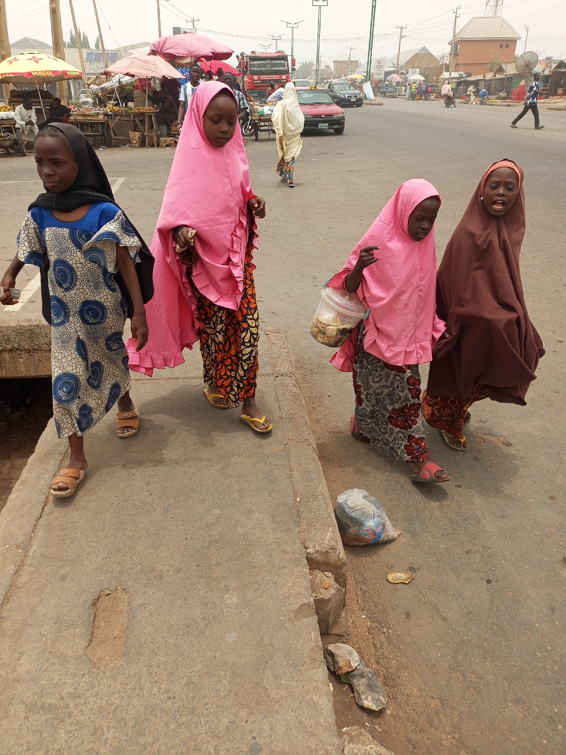 This photograph captures a bustling street scene in Tempure, Nigeria, featuring four young girls walking along a concrete curb during daytime. The sky appears hazy or overcast, diffusing the light.
In the foreground, from left to right, a young girl wearing a blue and white patterned dress and a black headscarf walks with her head slightly down, holding a small white item in her right hand. Next to her, another girl in a long pink headscarf and a vibrant orange and black patterned skirt also looks downwards. The third girl, similarly dressed in a pink headscarf and patterned skirt, carries a clear plastic bucket containing what appear to be baked goods or snacks, while pointing forward with her right hand. On the far right, the fourth girl, wearing a long brown head covering and patterned skirt, has her mouth open, suggesting she might be speaking or singing. All the girls are wearing sandals or flip-flops.
The background illustrates a busy urban environment. Numerous people are present, some walking, others tending to market stalls. Several market stalls are visible under colorful umbrellas, with one yellow umbrella prominently displaying the text "MAGGI." Goods are laid out on tables beneath these stalls. Vehicles, including a large red truck with "DAF" visible on its grille and a red sedan, navigate the asphalt road. Utility poles and distant buildings complete the setting, depicting a lively street in a West African town.