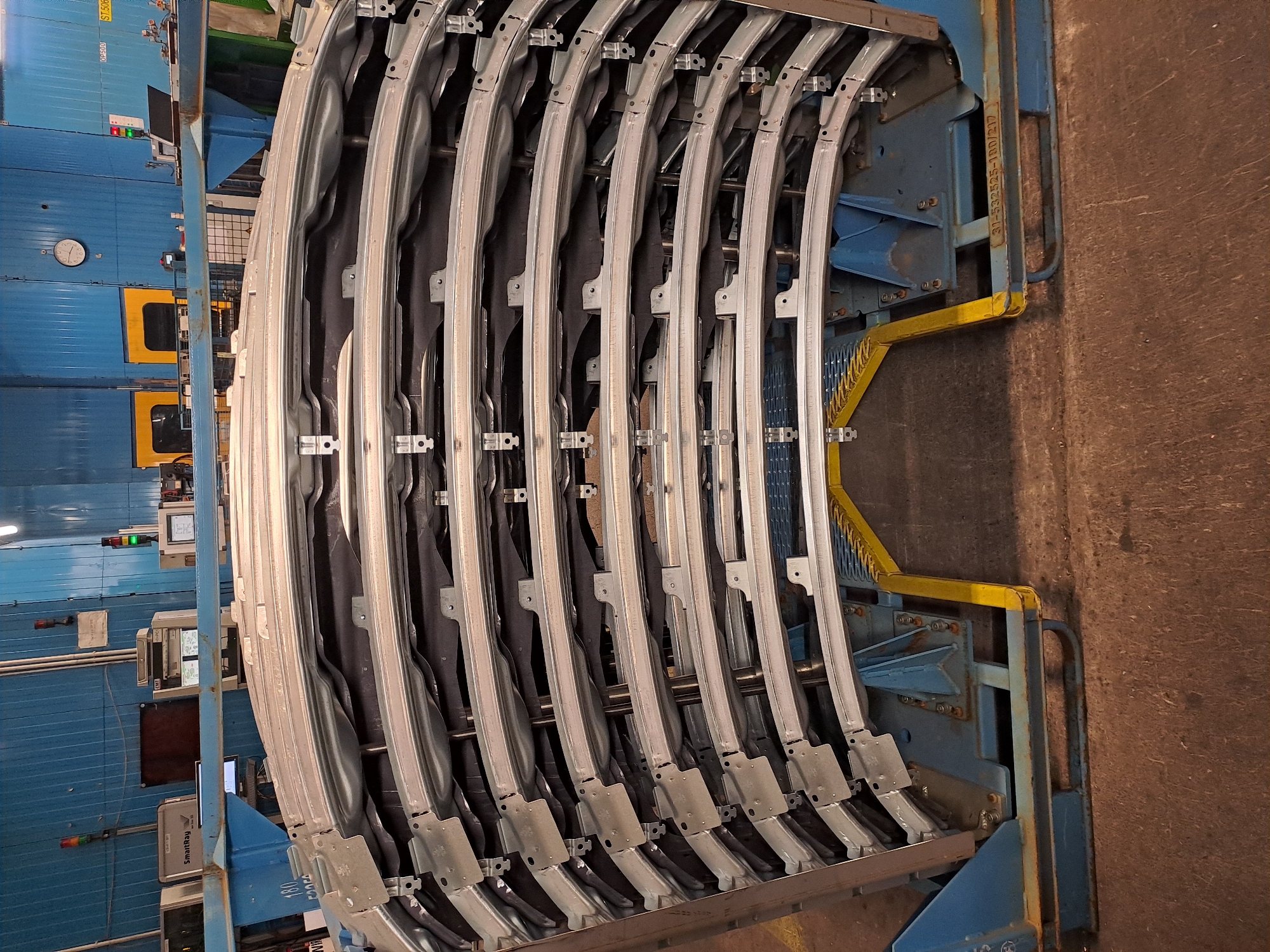 The image shows a metal rack filled with several curved, metal automotive components, possibly door frames or window frames, within an industrial setting. The rack is blue with yellow safety elements, and the components are arranged in a semi-circular pattern. The background includes a large blue industrial machine with monitoring equipment attached, as well as a brown industrial floor. There are no people visible in the image. The location, Haynrode, Germany, is not explicitly confirmed visually but implied from the context of the request.