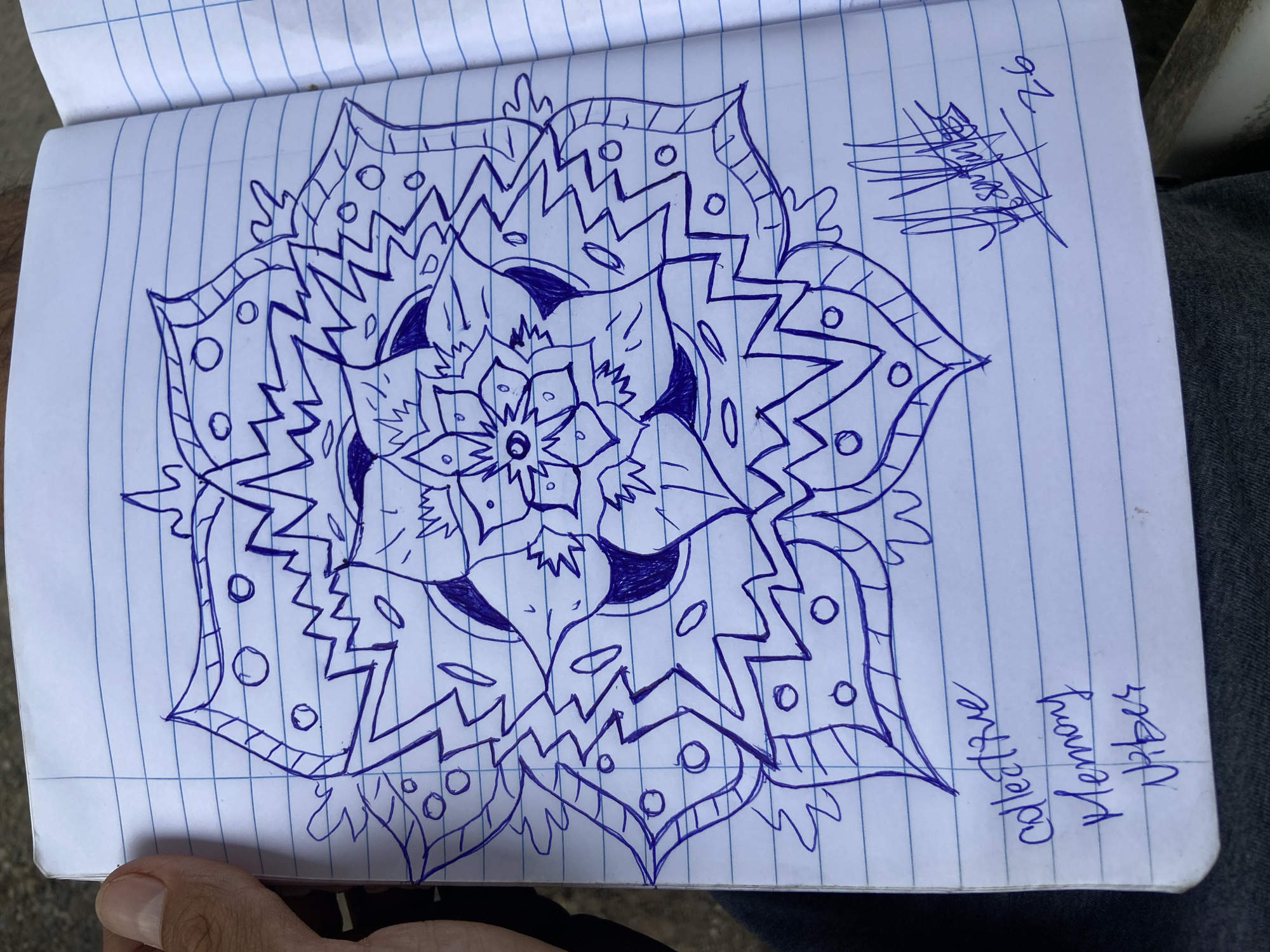 A notebook with lined paper displays a blue ink drawing of a mandala-like design. The drawing features a central floral pattern surrounded by layers of geometric shapes, lines, and circles. Text is visible on the right side of the page, including "Collective Memory Vibes" and a signature followed by "26". A hand is visible on the bottom left corner of the notebook. The notebook rests on a person's lap, wearing grey jeans. The location, Tinaquillo, Venezuela, is not visibly discernible from the image alone.