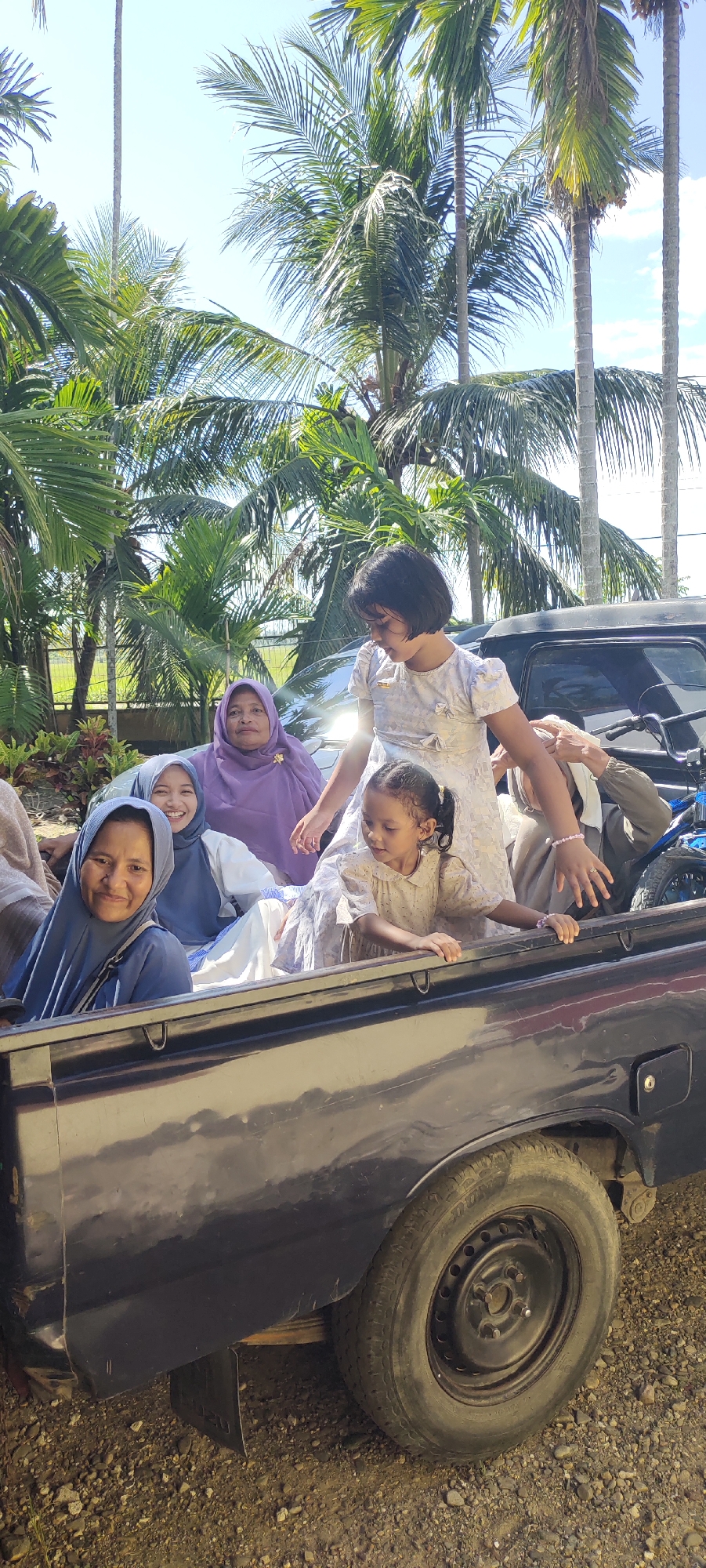 A group of people, including two young girls and several women wearing headscarves, are riding in the back of a black pickup truck. The environment includes palm trees and a sunny sky. The location appears to be an open area with other vehicles and possibly residential buildings visible in the background.