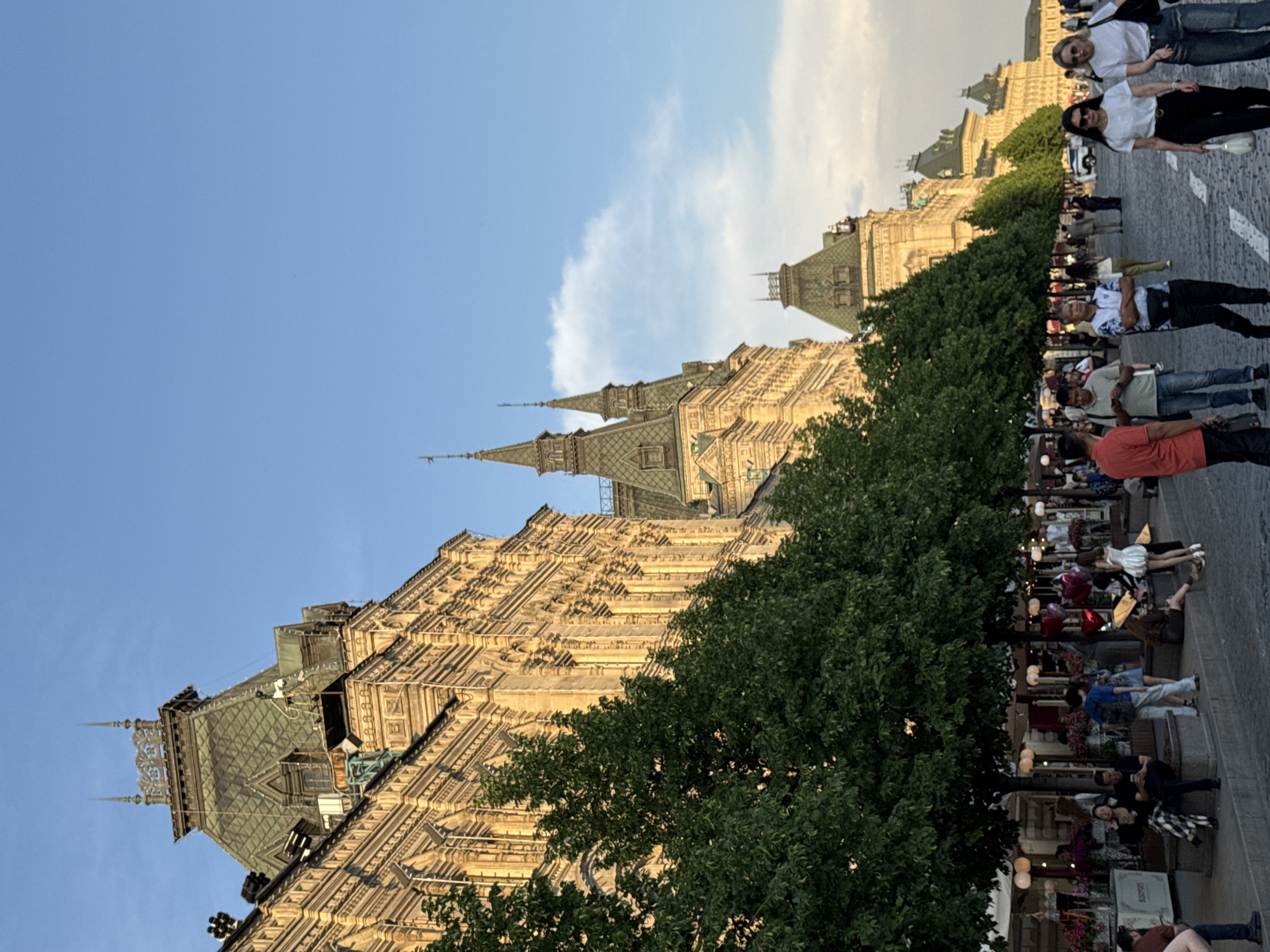 The image depicts a scene in Moscow, Russia, featuring the State Historical Museum. The building is ornate, with multiple towers and detailed facades. The sky is blue with scattered clouds. A line of trees runs along the side of the museum. People are walking along the cobblestone pavement in front of the museum. Tables and chairs of a sidewalk cafe are on the right side of the image.
