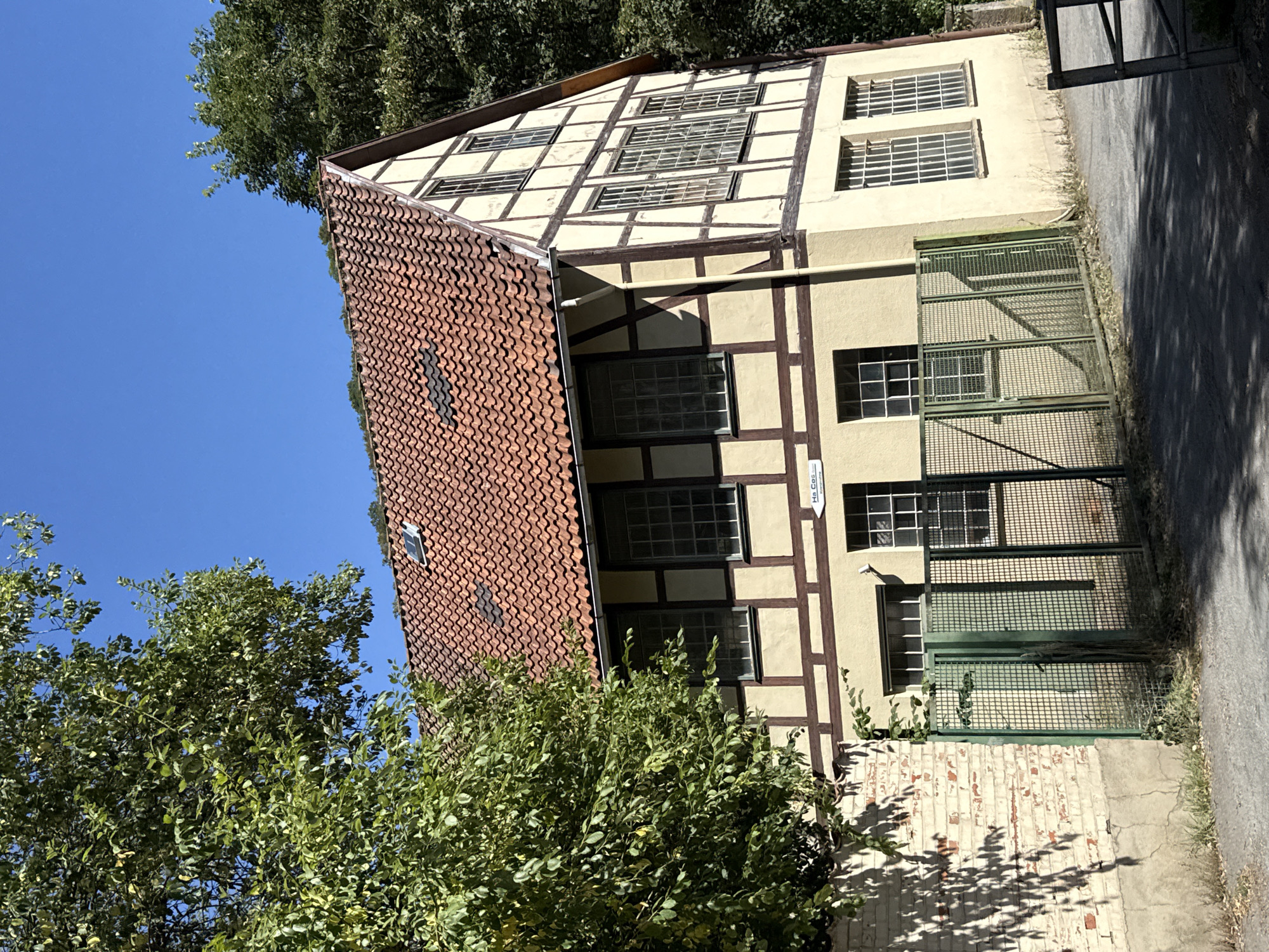 The image depicts a traditional German half-timbered house with a red tile roof, located at Schützenweg 1, in Bad Lauterberg im Harz, Germany. The house is painted yellow with brown timber framing and features numerous windows. In front of the house, there is a green metal fence and a paved area. A large, leafy tree partially obscures the left side of the building. The sky is clear and blue, suggesting a sunny day. A small white sign with black text, "Ha Cos," is visible on the exterior wall of the building, near the fence. The overall scene is one of a residential or perhaps an older commercial building in a German town.