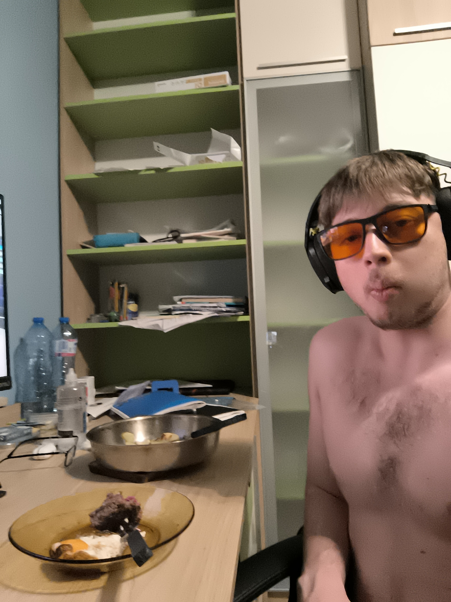 The image shows a shirtless man with headphones and orange-tinted glasses sitting at a desk. On the desk are a plate with food (possibly meat and fried egg), a frying pan, water bottles, a bottle of hand sanitizer, glasses, and papers. Behind the desk is a cabinet with shelves. The location is likely indoors, potentially an apartment or dorm room. There is no information in the image to determine the city of Sofia, Bulgaria.