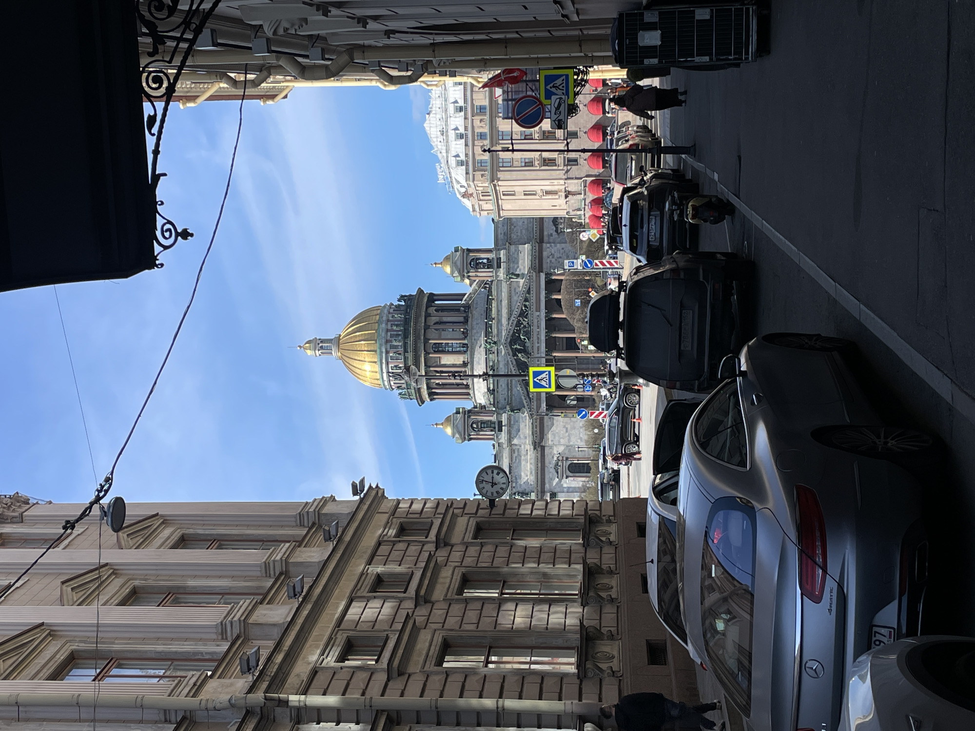 The image presents a sunlit street view in Saint Petersburg, Russia, framed by architectural structures on either side, leading the eye towards the iconic St. Isaac's Cathedral in the distance. The cathedral's prominent golden dome and classical facade dominate the central background under a clear, bright blue sky.
The scene is illuminated by natural light, suggesting midday, further supported by the visible round clock on the building to the right, which appears to show approximately 11:40. Buildings with ornate, classical architectural details line both sides of the street. The building on the left features a dark, decorative awning in the foreground. The building on the right showcases layered stonework, rectangular windows, and a series of external light fixtures.
The paved street below is lined with parked cars on the right. A silver Mercedes-Benz sedan with "4MATIC" badging is clearly visible, displaying a partially obscured Russian license plate ending in "97 RUS". Another car's license plate includes "921 M". A dark SUV with a roof rack is also parked further down the street. Several pedestrians are scattered along the pavement, one noticeable near the dark SUV.
Traffic and informational signs are visible along the street, including a yellow diamond-shaped pedestrian crossing sign, a red circular "No Stopping" or "No Entry" sign, and a blue circular "Direction to Follow" sign. The overall setting captures a blend of historical grandeur and everyday urban life.