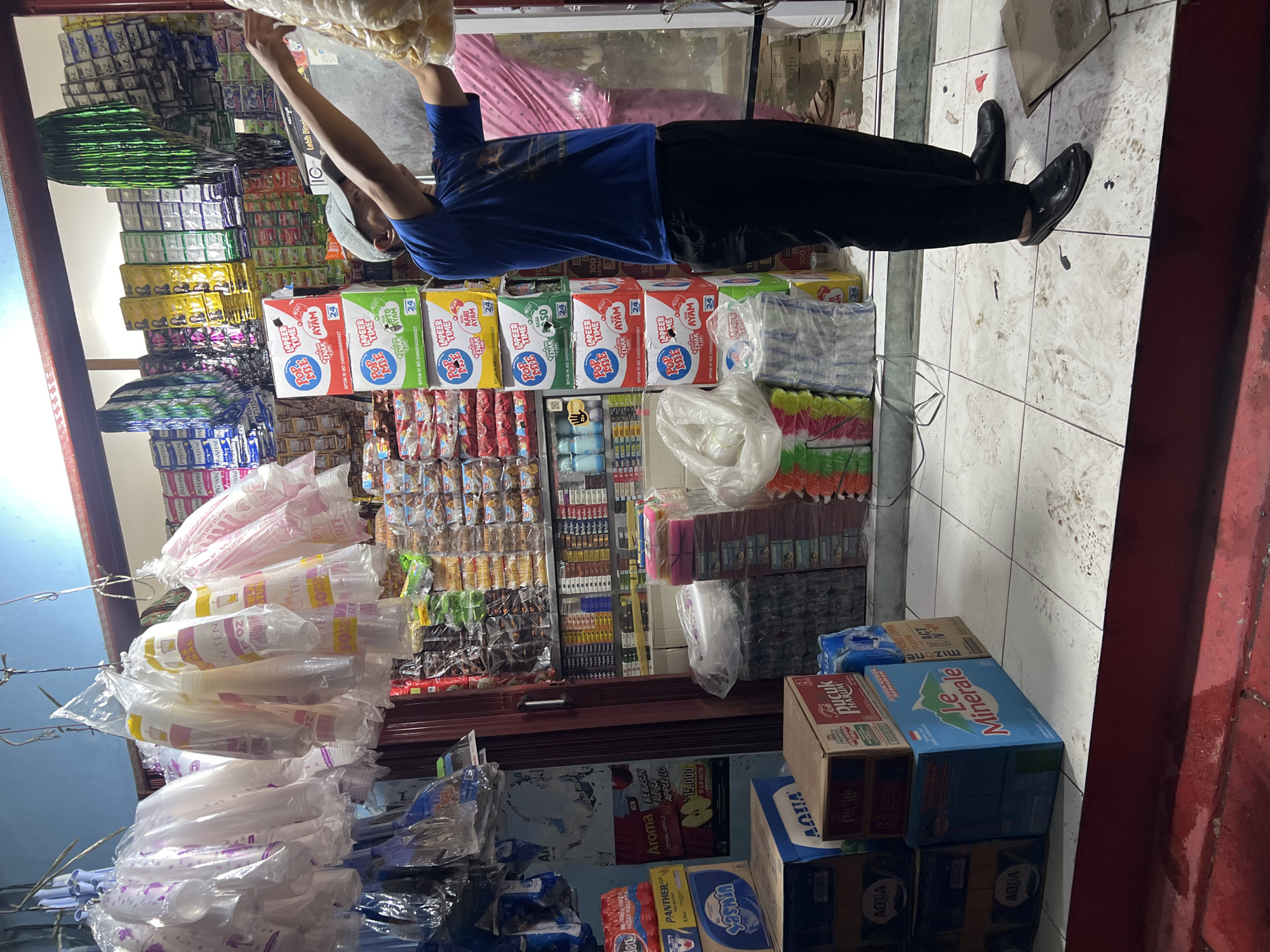 A man wearing a blue t-shirt and black pants stands in a small convenience store. He is reaching for a bag of goods hanging from the ceiling. The store is filled with shelves stocked with various products, including drinks, snacks, and household items. Plastic cups are hanging in the foreground. A woman in a pink outfit is visible in the background. The floor is tiled and appears somewhat dirty. The store seems to be located in Citeureup, Indonesia, as indicated by the visible product packaging.