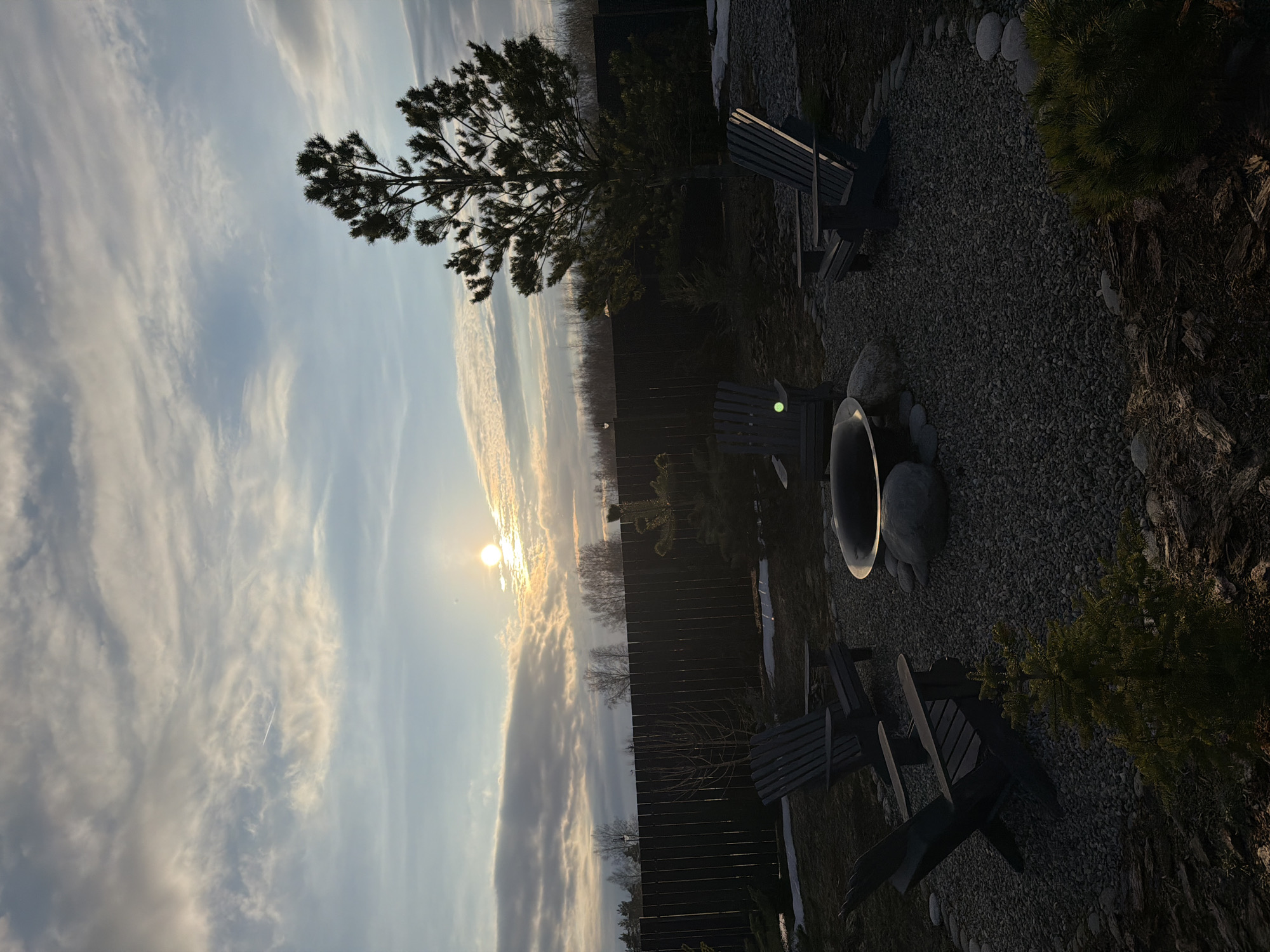 The image depicts an outdoor scene, likely a backyard or patio. The sky is visible with the sun shining through some clouds. On the ground, there's a fire pit surrounded by rocks and gravel. There are also several dark-colored Adirondack chairs scattered around the fire pit area. A wooden fence runs along the back of the scene, and there are some small trees and bushes. The image is rotated counter-clockwise. There is no evidence to suggest the location is in Ryazan, Russia.
