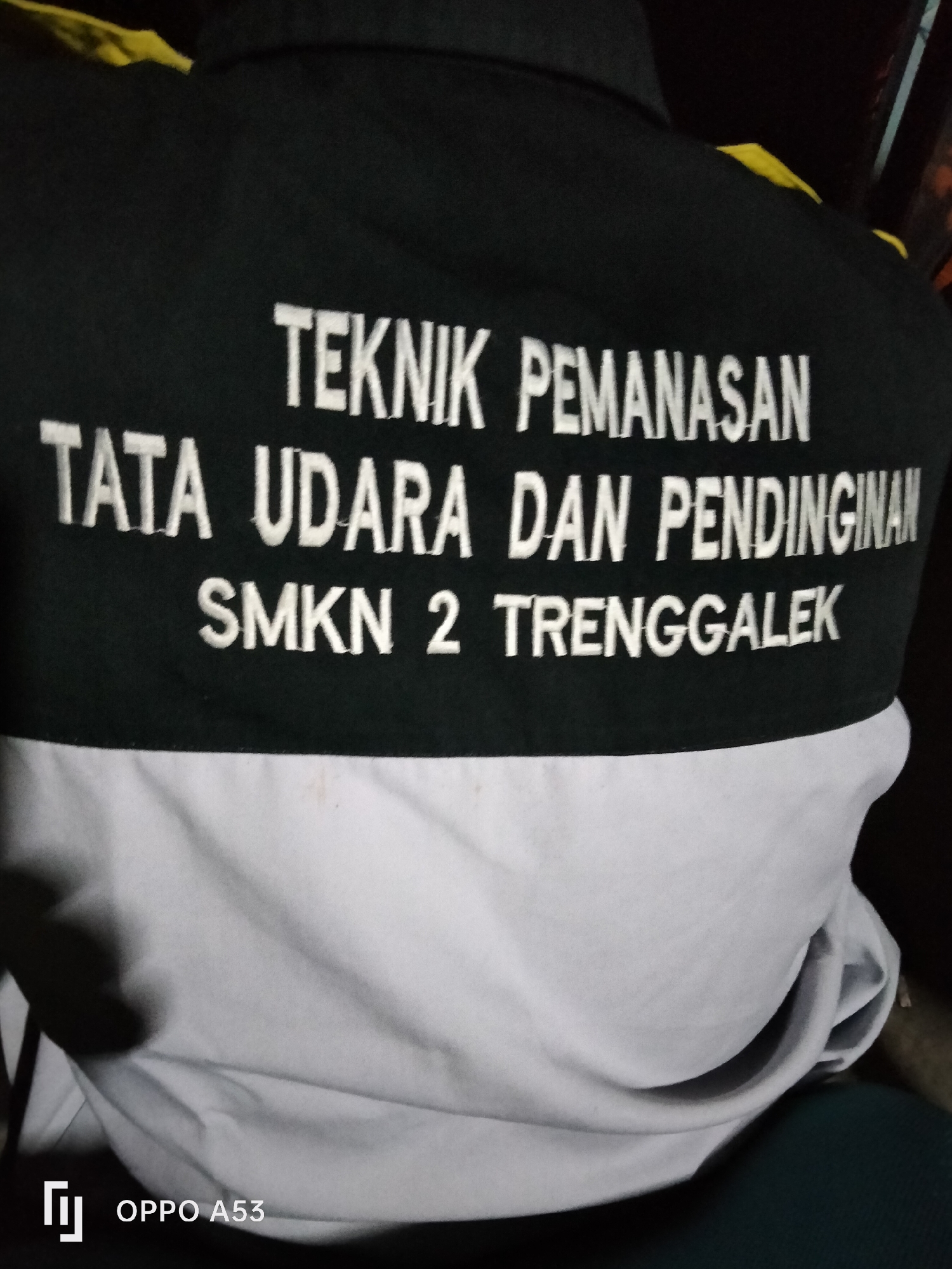 A close-up, slightly blurred, over-the-shoulder shot shows the back of a black and white shirt with white embroidered text. The top portion of the shirt is black, featuring yellow accents on the shoulders. The embroidered text on the black portion reads: "TEKNIK PEMANASAN TATA UDARA DAN PENDINGINAN SMKN 2 TRENGGALEK." The bottom portion of the shirt is white. In the bottom left corner, a logo and the text "OPPO A53" are visible. The background is dark and out of focus. The image is not located in Denpasar, Indonesia.