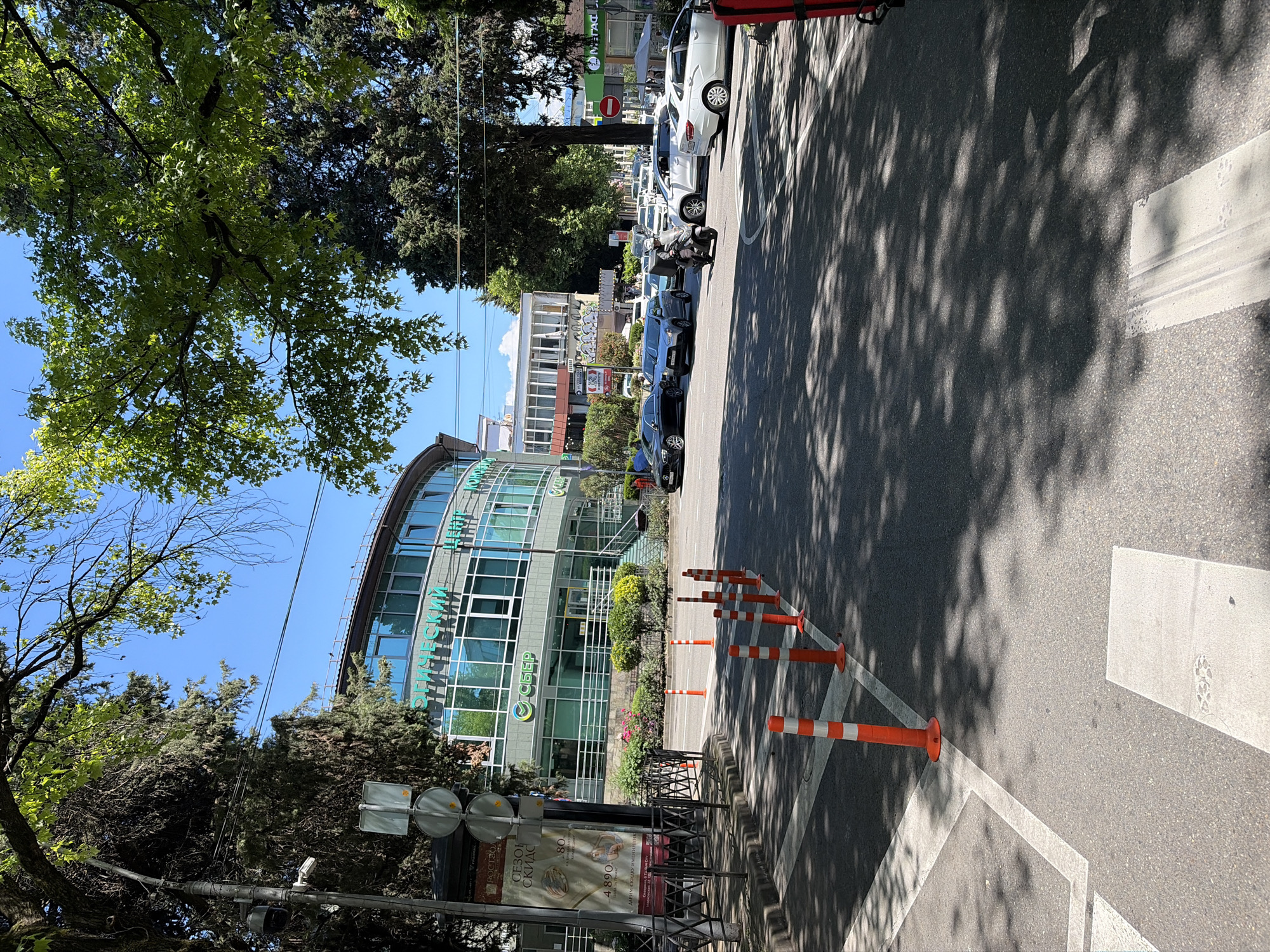 This image depicts a sunny urban street scene at an intersection in Sochi, Russia, specifically identified as the area around ul. Navaginskaya, 9D.
In the foreground, the asphalt road exhibits white painted markings, including a crosswalk on the left and a stop line or parking indication on the right. A series of orange and white striped traffic bollards are arranged along the edge of the road, separating it from the sidewalk on the left. Dappled shadows from overhead trees are cast across the asphalt.
To the left, a metal fence runs alongside a sidewalk, behind which a street lamp post with attached CCTV cameras stands. An advertising stand displays a poster with Russian text, including "СЕЗОН СКИДОК" (SEASON OF DISCOUNTS), "РОСТЭЛ" (ROSTEL), and a price of "4 890". Lush green foliage and dense trees line the left side of the street.
The midground features an active intersection with multiple vehicles. A person wearing a helmet and a dark jacket rides a grey scooter or moped, positioned centrally on the road. Various cars are present, including a black sedan, a grey SUV, a white SUV, and a white sedan, all appearing to be stopped or moving slowly through the intersection. A red circular "No Entry" sign with a white horizontal bar is visible on the right side of the road.
Dominating the background is a large, curved multi-story building with a modern design, featuring numerous windows with a greenish-blue tint and a light grey facade. Prominently displayed on this building are the green logos and text "СБЕР" (Sber) and "ОГИЧЕСКИЙ ЦЕНТР" (OLOGICHESKY TSENTR, likely an ophthalmological or similar medical center). Further in the background, other commercial or residential buildings with white or light-colored facades are visible, along with additional trees. A red logo for "ПЯТЕРОЧКА" (Pyaterochka), a Russian supermarket chain, is partially visible on the far right. The sky above is clear and bright blue with minimal cloud cover.