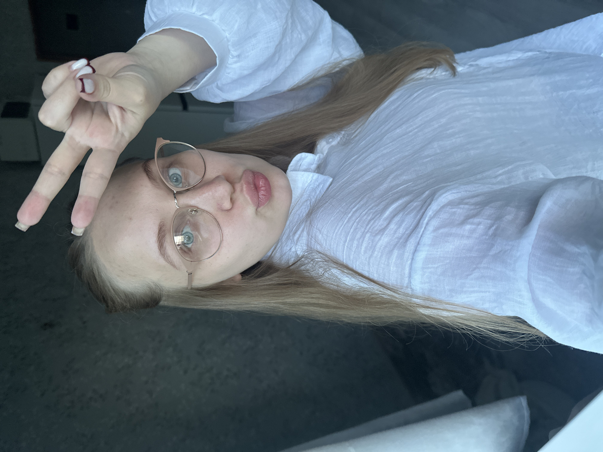 A light-skinned young woman is angled with her head toward the bottom left of the frame, wearing round, gold-rimmed glasses and a white, collared shirt. She is holding up her left hand, making a peace sign next to her forehead. Her nails are painted white with a maroon accent at the base. Her long, light brown hair falls over her left shoulder and onto the white shirt. The background is out of focus, but appears to be a dark surface. The location is not discernible from the image. The city of Chaikovsky, Russia, cannot be confirmed based on the image.