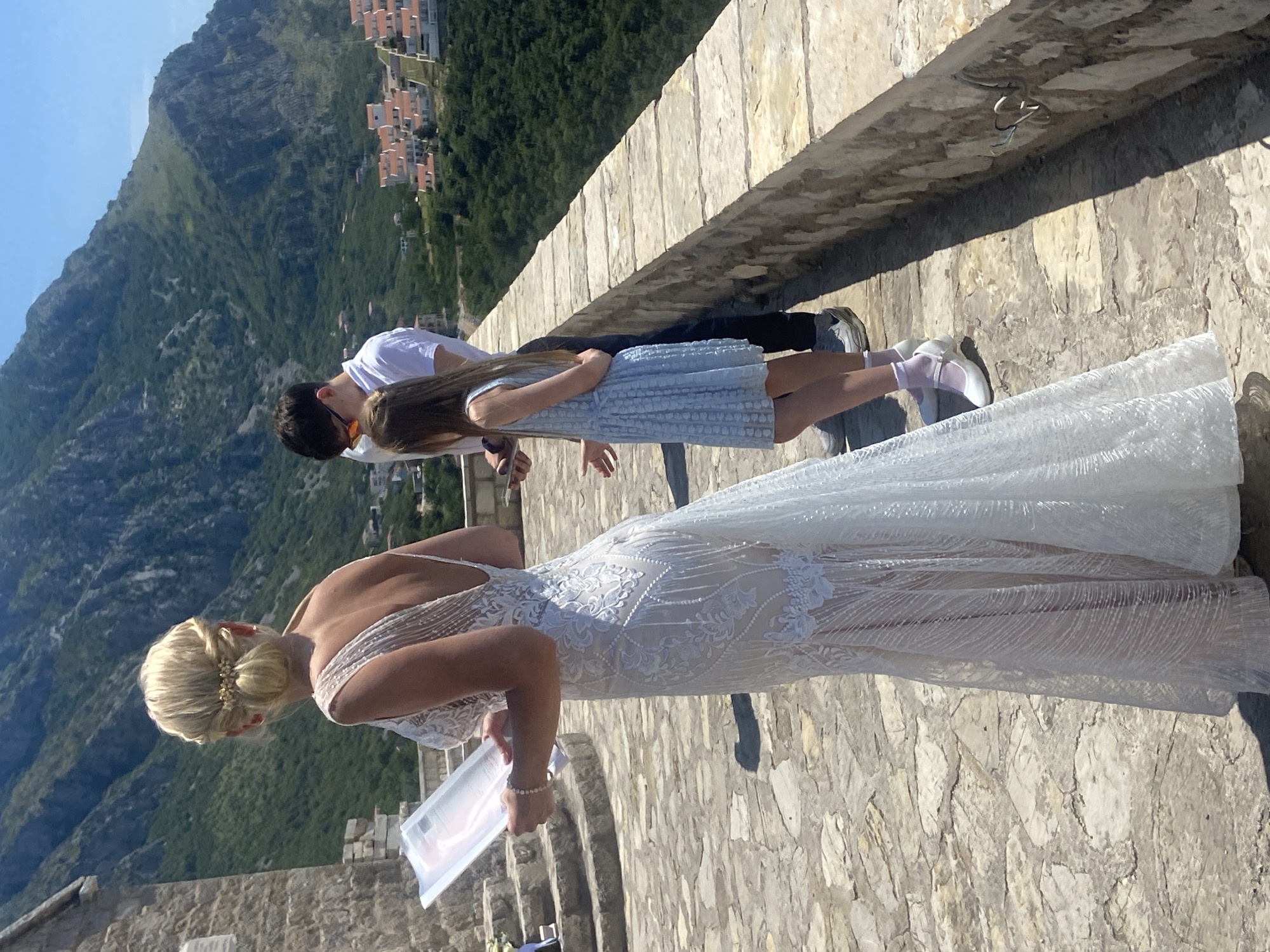 A woman in a white lace wedding dress stands holding a document and looking down. She has blonde hair styled in an updo with a decorative clip. To her right, a young girl in a light blue dress stands looking down, and behind her, a young man in a white t-shirt with sunglasses on his head also looks down. The group is on a stone walkway with a stone wall to their right. In the background, there are lush green mountains with buildings clustered on a hillside. The scene appears to be an outdoor wedding ceremony in Sveti Stefan, Montenegro, overlooking the Adriatic coast.