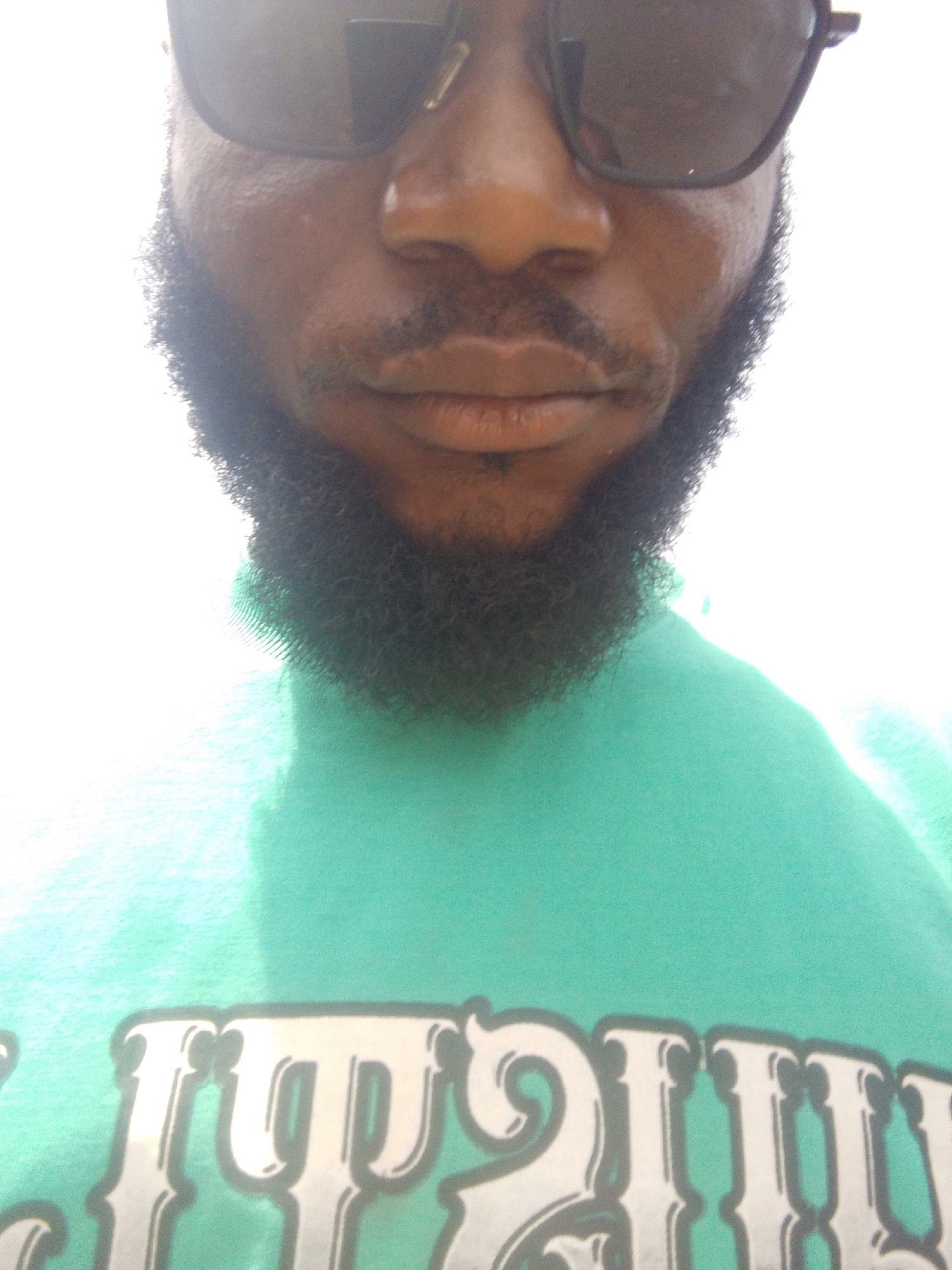 An individual with a dark complexion, wearing sunglasses and a beard, is positioned in the center of the frame. The person is wearing a green shirt. The background is predominantly bright white. The location is not determinable to be Lokoja, Nigeria from the given image.