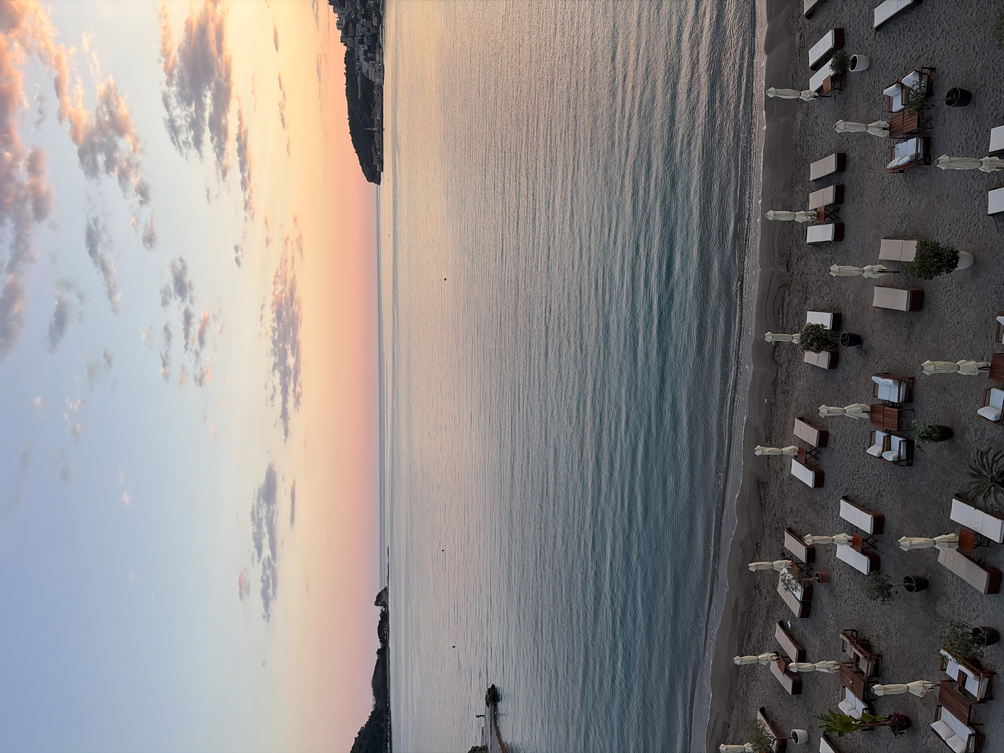 The image shows a beach in Budva, Montenegro from an elevated perspective, likely taken during sunset. The sky displays a gradient of colors from blue to orange. The calm sea reflects the sky's colors. Along the beach, there are numerous sun loungers with white umbrellas arranged in neat rows. Some of the seating areas have small tables and potted plants. The beach appears to be mostly empty. The far side of the bay is outlined by a dark landmass, possibly a peninsula, with buildings visible along the coastline.