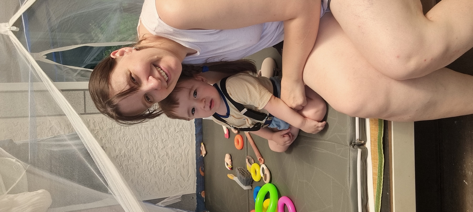 A medium, indoor shot depicts a woman and a child within a tented structure, potentially a play area. The woman is fair-skinned with brown hair, wearing a white top, and she smiles towards the camera. A small child with brown hair, wearing a tan shirt and suspenders, is positioned on the floor. On the floor are various toys with rings, handles, and a brush. The tent is enclosed with a net material and a white interior wall is partially visible. The location is Dunnae-myeon, South Korea.