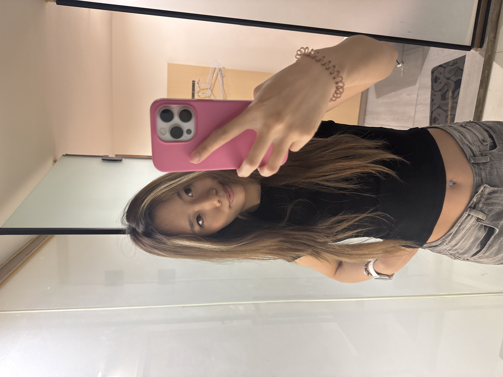 The image captures a mirror selfie of a young Asian woman, oriented 90 degrees counter-clockwise. She is holding a pink smartphone with a multi-lens camera system in her right hand, using it to take the photo. Her long, straight brown hair, featuring lighter highlights, cascades over her shoulders. She is wearing a black short-sleeved crop top that exposes her midriff, where a belly button piercing with a small, light blue gem is visible. Her lower body is dressed in light-colored, possibly grey, jeans. On her left wrist, she wears a white Apple Watch, and on her right wrist, a coiled, spiral hair tie or bracelet.
The woman is looking directly at the camera with a slight, casual smile, conveying a relaxed demeanor. The scene appears to be an indoor setting, likely a fitting room or a modern room with a large mirror. The background, visible in the mirror's reflection, consists of light-colored walls. In the upper left background, a faint reflection of what might be a white metal clothing rack or similar furniture is discernable. A section of floor with a grey and white geometric pattern is visible behind her. The lighting is even, suggesting an interior artificial light source. No discernible text is present in the image. The provided location context for the image is Yuzhong District, China.