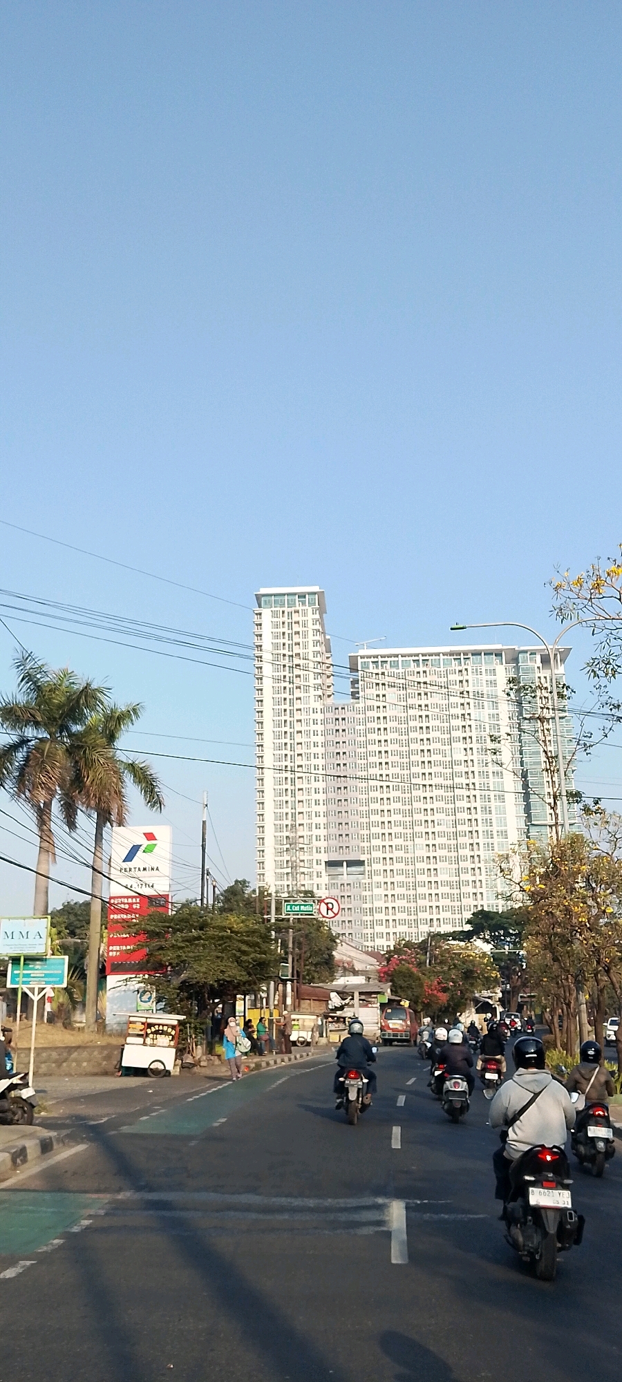 This is a daytime, eye-level, full-frame shot of a street scene in Bekasi, Indonesia, looking north. The sky is clear and blue. In the background, two tall, modern, white apartment buildings with many windows dominate the skyline. To the left, two palm trees are visible in front of a Pertamina gas station sign. The street is paved and has multiple lanes. Several motorcycles, some with riders, are traveling on the road. On the sidewalk, a few pedestrians can be seen, along with small food carts. A street sign indicates "Jl. Cut Mutia" with a "no U-turn" symbol. The overall impression is a busy urban environment.