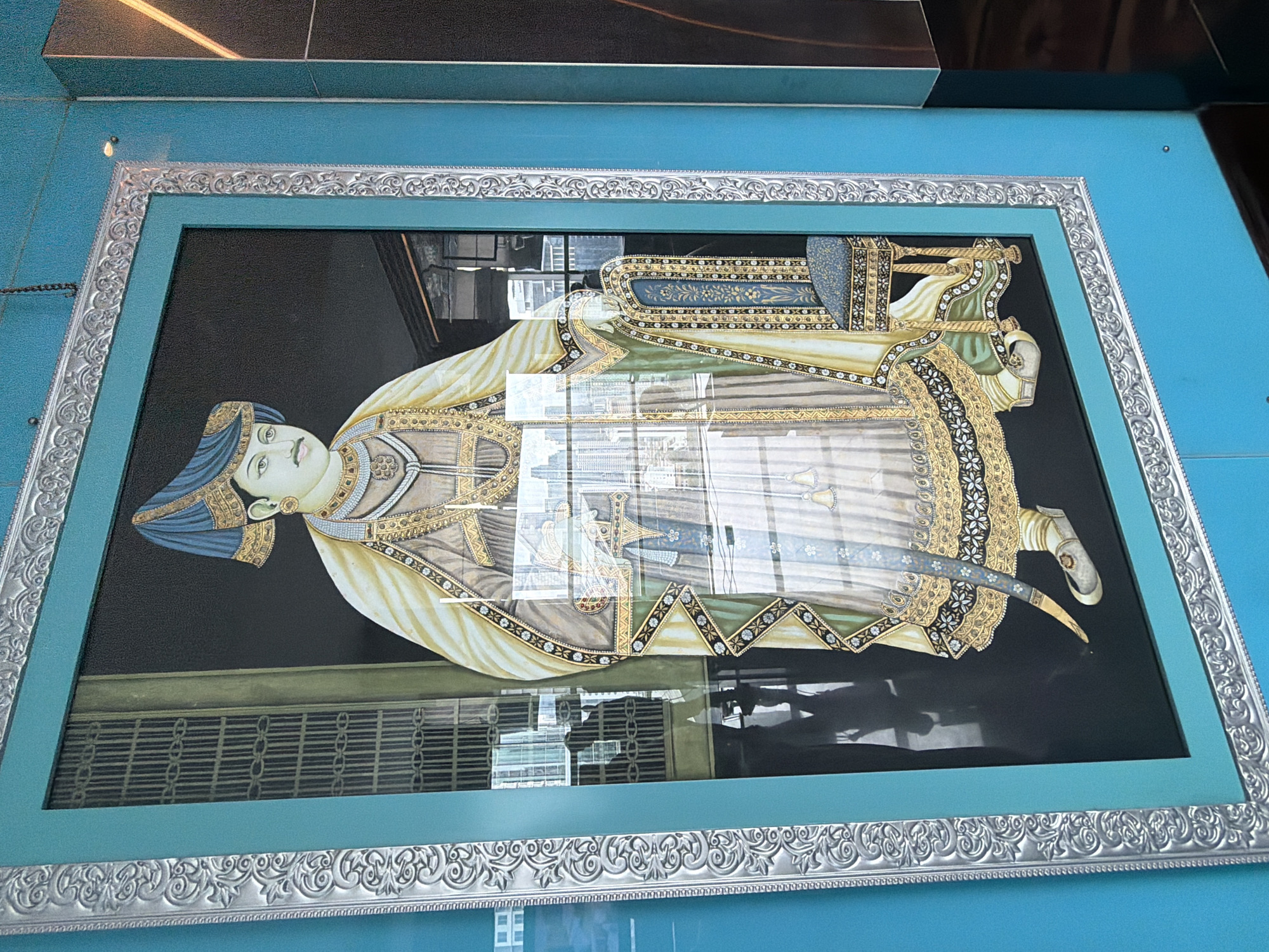 The image shows a framed portrait of an Indian man in traditional clothing, possibly a royal figure, displayed against a bright blue background. The frame is ornate, with silver-toned detailing. The painting features a man with a mustache, wearing a blue turban and a heavily embellished robe. The background of the portrait is black. Reflections are visible on the surface of the portrait, obscuring some details of the artwork and indicating the presence of a glass or plastic cover. A second frame is behind the first. The image was taken indoors at South Jakarta, Indonesia.
