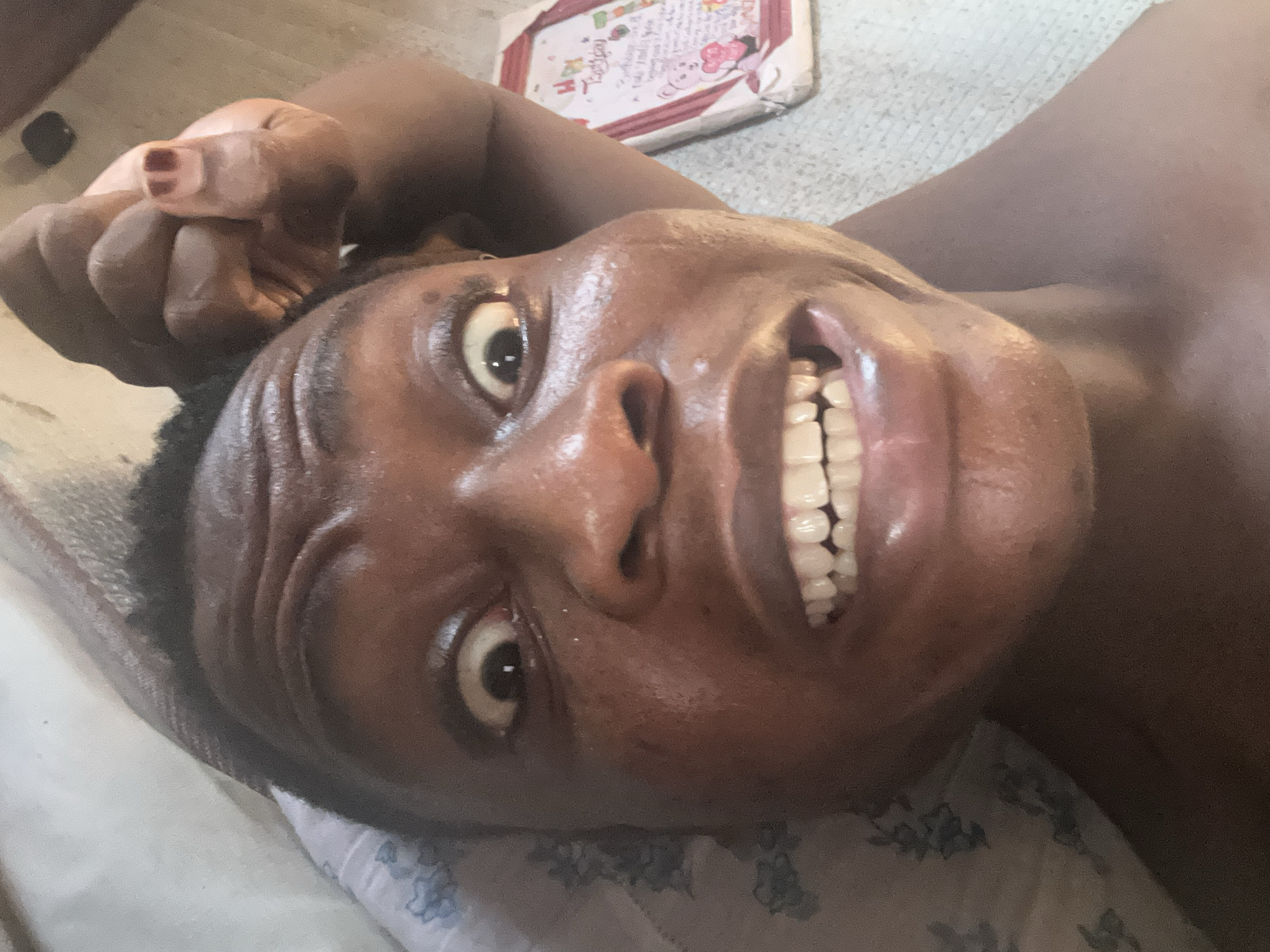 A close-up, angled view depicts a person lying down. The person has dark skin, visible wrinkles on their forehead, and shows a full, wide smile revealing bright white teeth. The person's right hand is clenched near their head, with a glimpse of what appears to be reddish-brown nail polish. The background includes a partially visible pillow with a blue floral pattern and what seems to be a small, decorated board or box. The location is unknown.