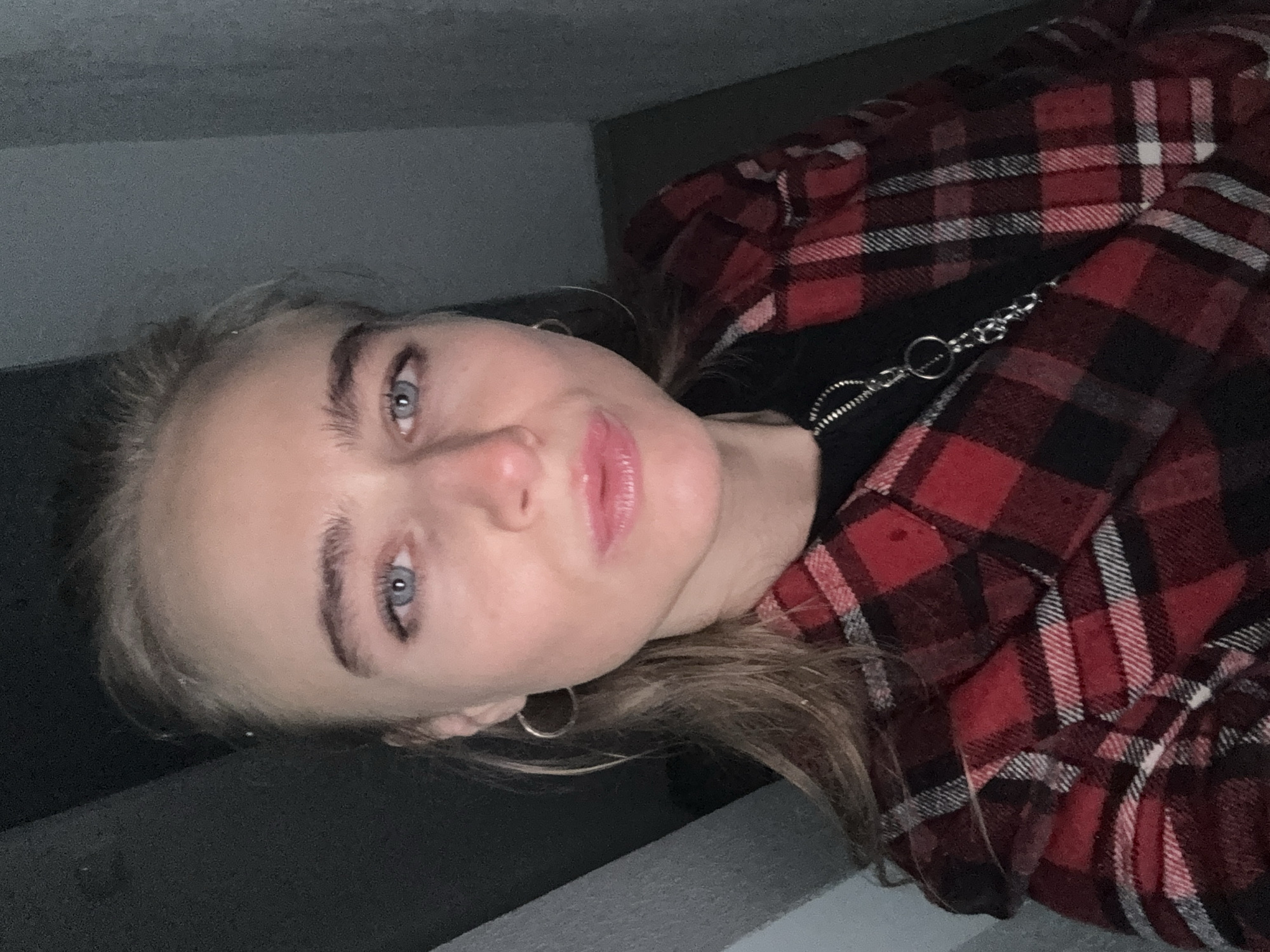 The image features a close-up, rotated portrait of a young woman. Her head is tilted, and she is looking directly into the camera. She has light-colored hair, possibly blonde or light brown, and striking blue eyes. Her eyebrows are well-defined, and her lips appear glossy, with a light pink hue. Small, silver hoop earrings are visible on her ears.
She is dressed in a red and black plaid flannel shirt or jacket worn over a black top. A silver chain-link accessory is visible around her neck, resting on the black top. The background is indistinct, appearing to be an indoor setting with dark, plain walls or a ceiling.
The lighting in the image is somewhat dim and uneven, suggesting an indoor environment possibly illuminated by artificial light, or taken during low light conditions. Her expression is neutral with a soft, direct gaze. There is no discernible activity taking place other than posing for the photo. No text is visible in the image.