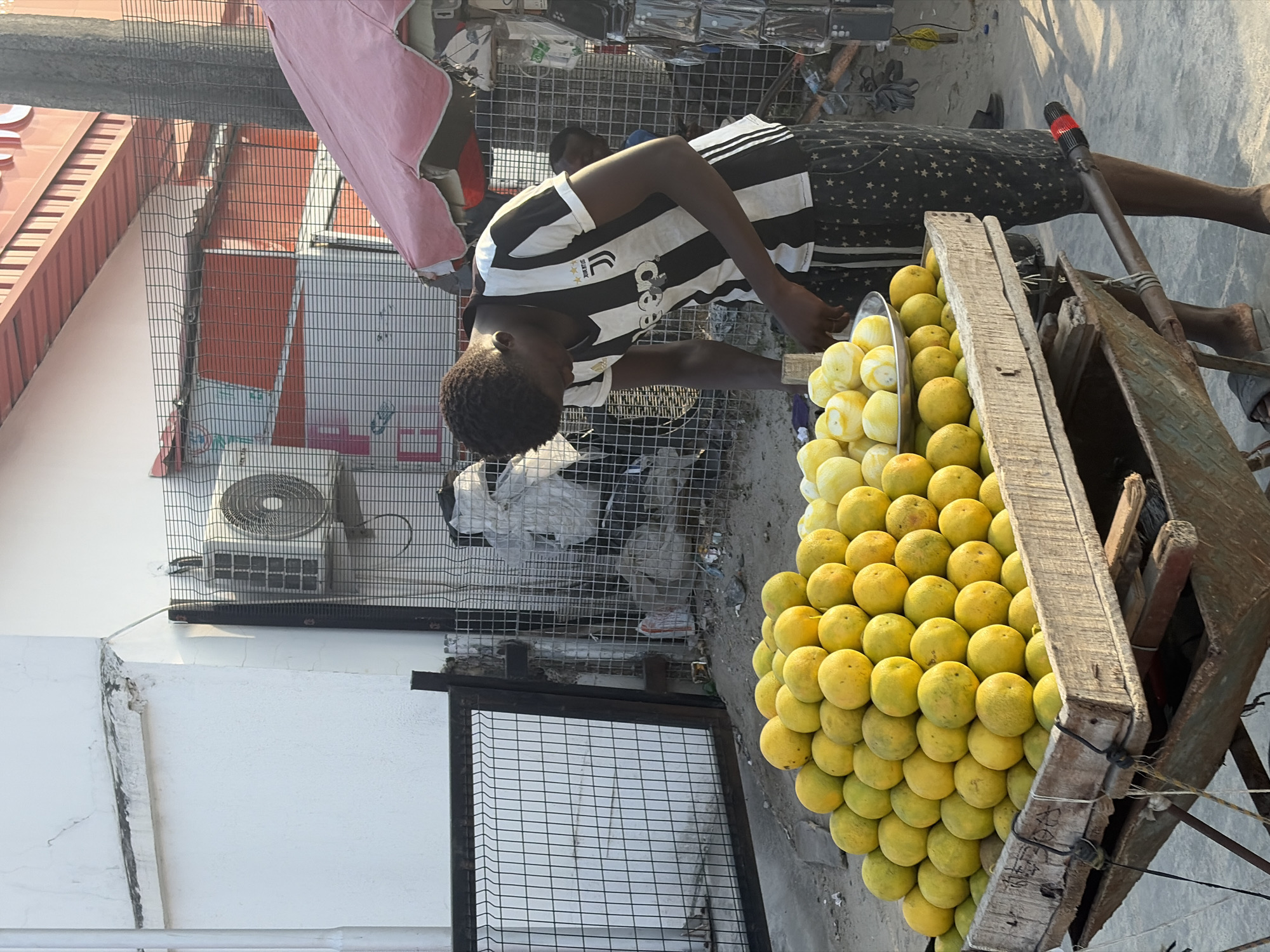 In Igboefon, Nigeria, a young man in a black and white striped jersey stands behind a pushcart laden with yellow citrus fruits, likely oranges or grapefruits. The cart is constructed from weathered wood and metal. Behind the vendor, a metal cage fence runs along the sidewalk. An air conditioning unit is mounted on a white wall behind the fence. The street is paved, and a pink cloth is partially visible overhead. The scene depicts a street vendor selling fruit.