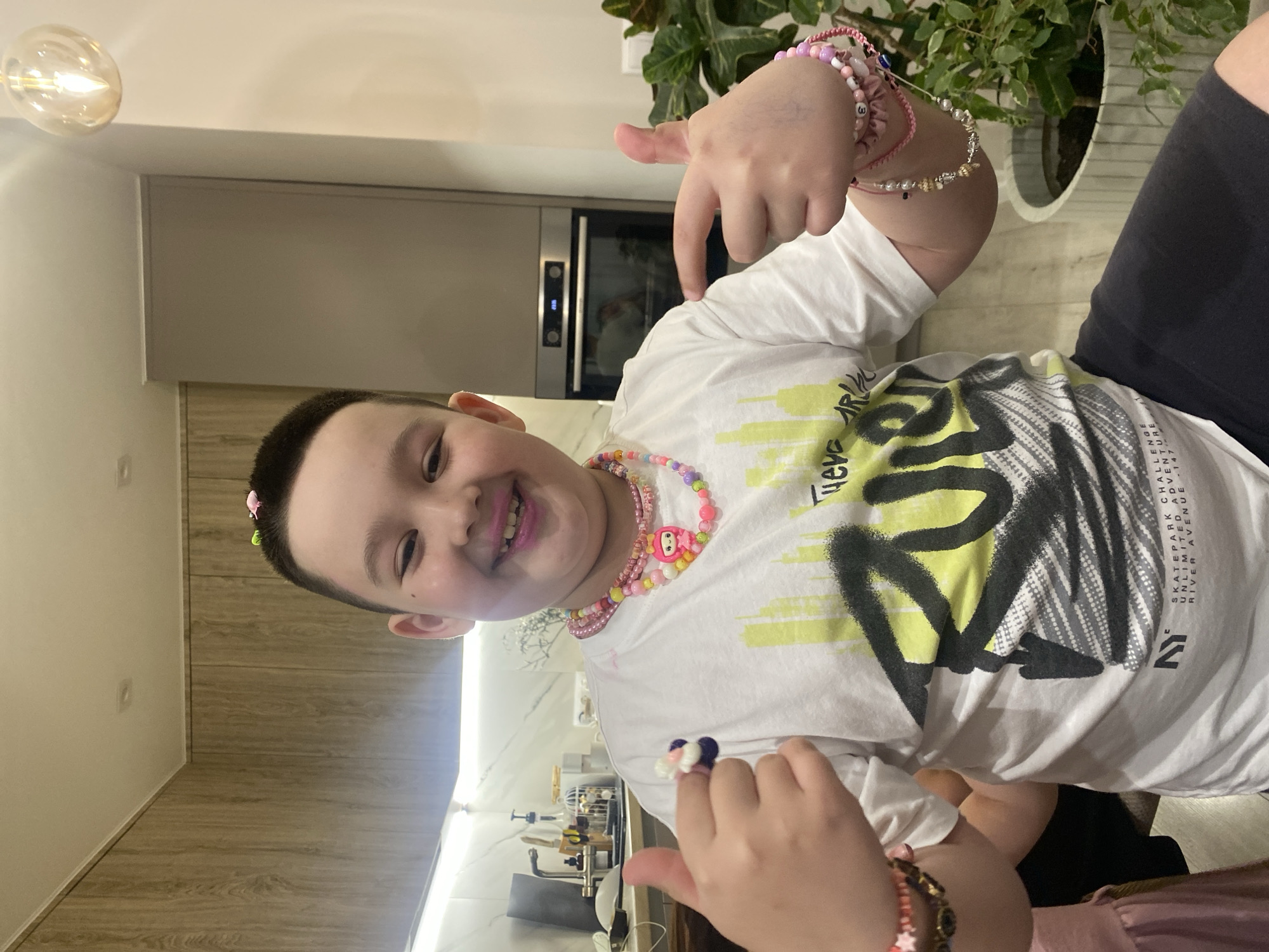 A young boy is posing indoors. He is wearing a white t-shirt with a yellow and black graphic design, two colorful beaded necklaces, and several bracelets on his wrists. His hair is dark and short, and he has small, decorative hair clips. The background shows a modern kitchen with light-colored cabinets and countertops, an oven built into the cabinetry, and a houseplant in the background. The location in the city of Izhevsk, Russia is not evident from the image.