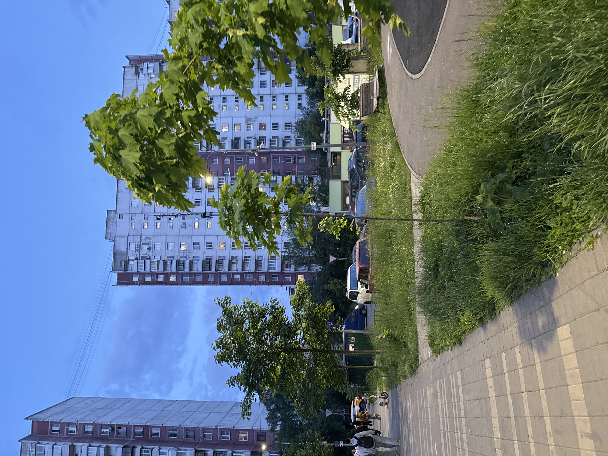 The image captures an urban or suburban street scene during dusk or early evening, characterized by the deep blue sky and illuminated windows in apartment buildings. The perspective is from ground level, looking down a paved sidewalk and a grassy strip alongside.
In the background, several multi-story apartment buildings dominate the skyline. They are block-style structures, some with lighter upper sections and darker, possibly red-brown, lower sections. Many windows are lit, indicating the time of day. Overhead, power lines or communication cables stretch between buildings.
The foreground features a grey, block-paved sidewalk extending into the distance on the right, bordered by a wide strip of lush green grass. Parallel to the grass and sidewalk, a darker, curved paved path (possibly a bike or jogging path) is visible. A wooden bench is partially seen next to this curved path.
Numerous green-leafed trees are scattered throughout the scene, with some prominent in the foreground and middle ground, providing a canopy effect. A lit streetlamp stands near one of the trees, casting light on the surroundings.
Along the grassy strip, several parked vehicles are visible, including a distinctive white and orange-brown van or truck and other cars. A few indistinct figures of people are seen walking on the sidewalk in the middle distance, one possibly with a scooter.
On the partial view of the wooden bench, some faint Cyrillic text is discernable, with the word "ЧАСТИ" (chasti) being somewhat visible, suggesting a location in a Russian-speaking country. The overall mood is peaceful and ordinary, reflecting typical evening activity in a residential area.