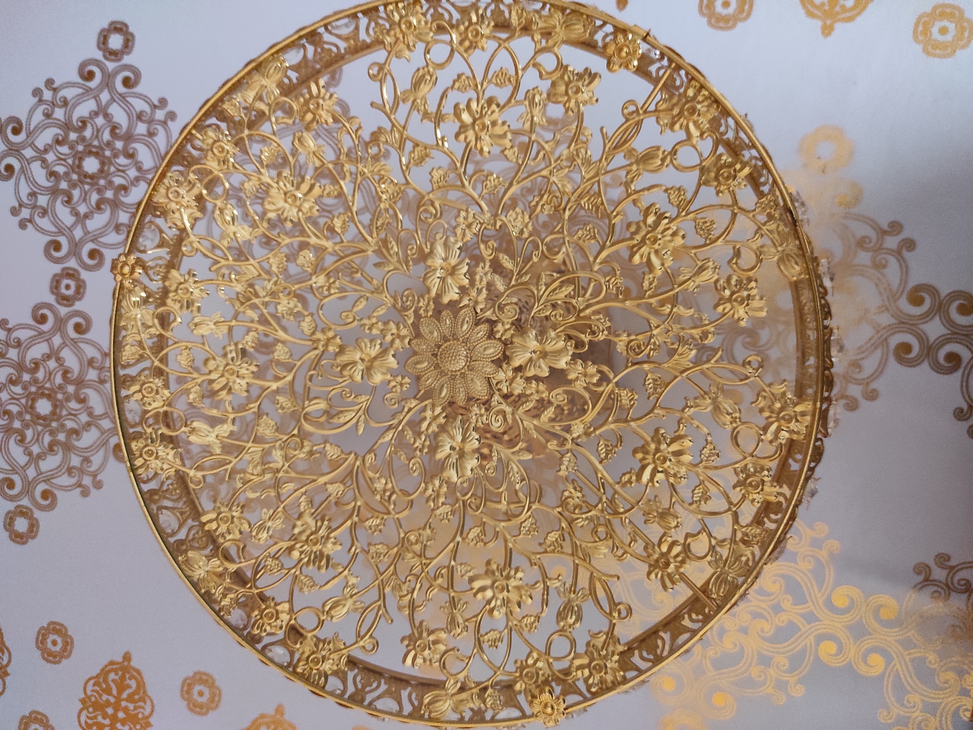 A close-up, high-angle shot captures an ornate, gold-colored metal ceiling fixture, possibly a chandelier or a decorative medallion. The fixture is circular and intricately designed with floral motifs and swirling vine-like patterns. The metal is a bright, polished gold, reflecting light and creating a sense of opulence. The fixture appears to be suspended from a white ceiling that is decorated with subtle, golden patterned wallpaper, featuring swirling arabesque designs and small floral elements. The lighting in the room is soft, casting faint shadows of the fixture onto the ceiling. The overall impression is one of grandeur and classical architectural detail. This ornate ceiling fixture is located in Khiva, Uzbekistan, likely within one of its historical madrasahs or palaces, such as the Tash Hauli Palace or the Muhammad Aminkhan Madrasah, known for their elaborate decorative elements.
