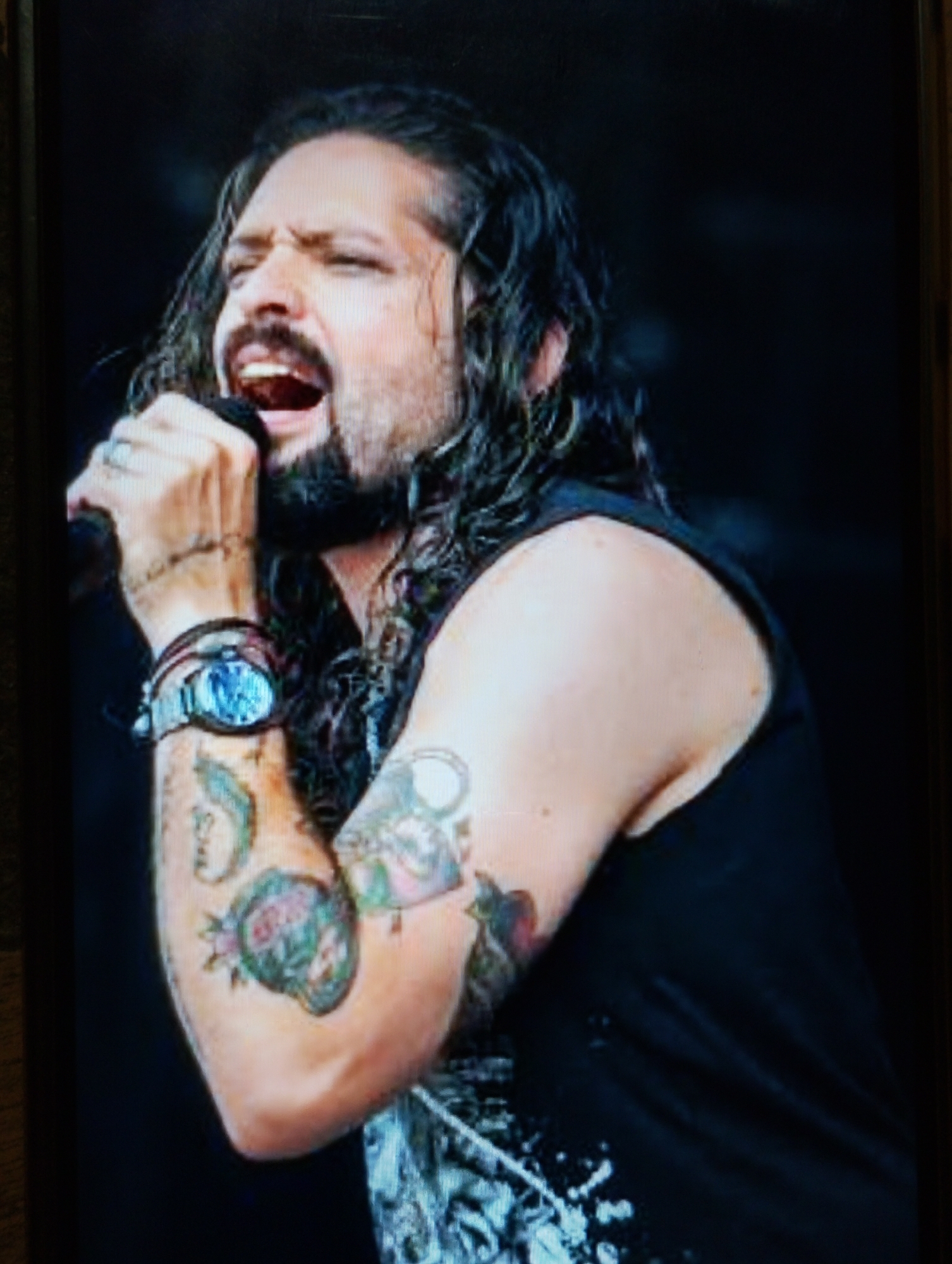 A man with long, dark, curly hair, a beard, and mustache is singing into a microphone. He is wearing a black sleeveless shirt, a watch on his left wrist, and a ring on his left ring finger. His left arm is covered in tattoos. The background is dark and out of focus. The image appears to be a photograph taken during a live performance.