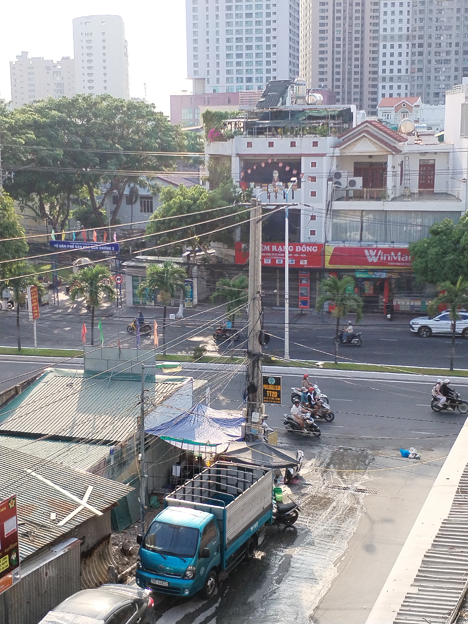 The image depicts an urban scene in Nha Trang Ward, Vietnam. A blue truck with license plate "79C-14802" is parked on a wet street. Near the truck, a gray car is parked as well. Debris and standing water are visible. Above the street, a WinMart convenience store can be seen. The street is fairly busy with scooter traffic. Power lines cross the frame, and apartment buildings are visible in the background.