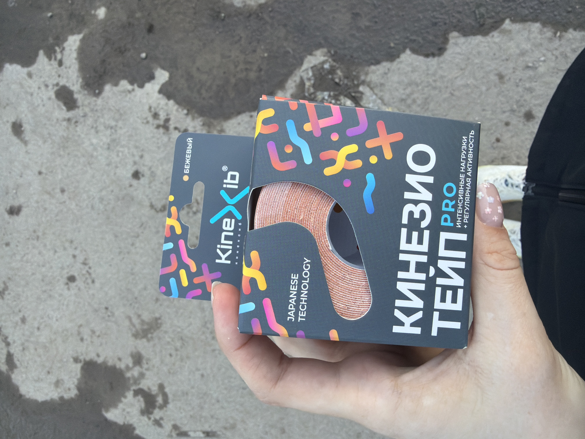 Here is a detailed description of the image:
A person is holding two boxes of Kinesio tape in their hand. The tape is beige and the boxes are gray with colorful geometric designs. "Kinexib" is printed on the left box and "Kinesio Tape Pro" in Russian on the right. The tape is indicated as "Japanese Technology". The background is a gray, wet concrete sidewalk. Based on the language and the context of the image, it is likely that this photo was taken in Russia, and specifically Berezniki is likely as the Kinexib brand is heavily marketed there.