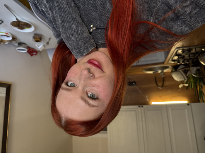 The image depicts a female individual taking a selfie in a residential interior, specifically a combined kitchen and dining area in Rostov-on-Don, Russia. The woman, positioned in the foreground, has long, bright red hair, fair skin with freckles, green eyes, a septum piercing, and red lipstick. She is wearing a grey, textured long-sleeved top and a necklace with a star charm, looking directly at the camera with a slight smile, her left hand resting near her chin.
The background shows a functional kitchen area to the left, featuring white upper cabinets with black handles, a light-colored wooden countertop, a stainless steel sink with a gooseneck faucet, and a four-burner gas stovetop with two pots. Two linear under-cabinet lights illuminate the workspace. A green plant in a white pot is visible on the countertop. To the right of the subject, a white dining table holds various food items, including a white oval plate with dark food and a green garnish, multiple small white bowls containing snacks, a glass jar with green contents, a paper napkin dispenser, and a small pastry in a clear container. A dark-framed picture hangs on a light-colored wall in the far right background.