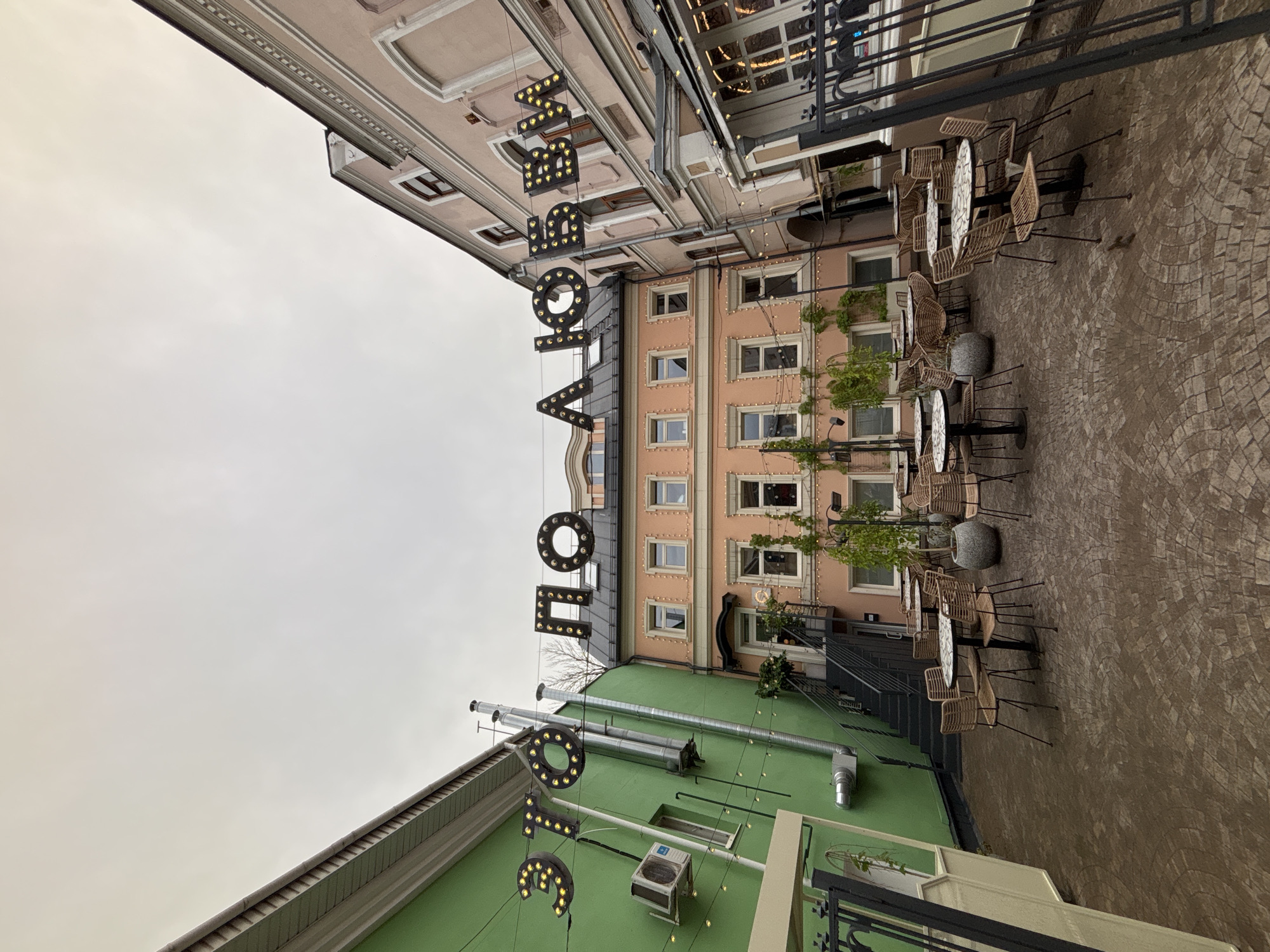 This media file is an exterior shot of a building with an outdoor seating area. The scene is a courtyard with cobblestone paving and several tables and chairs. The buildings surrounding the courtyard have a mix of architectural styles, with some featuring ornate detailing and others having a more modern facade. The lighting suggests it might be daytime, but the sky is overcast, giving a muted and somewhat somber feel to the image. There are no people visible, and the activity appears to be a dormant cafe or restaurant.
Notable details include illuminated signs hanging from the buildings. One sign appears to spell out "N O V O" with individual letters illuminated by small bulbs. Another sign below it also has illuminated letters. The buildings are decorated with strings of small lights, and there are potted plants and some greenery adding a touch of life to the scene. The presence of an air conditioning unit on the green-colored section of the building indicates it's a habitable structure. The cobblestone pavement suggests an older urban setting.
The location is confirmed as Nizhny Novgorod, Russia. The overall impression is a somewhat cool, potentially damp day in an urban European courtyard, likely during off-hours or perhaps before opening.
