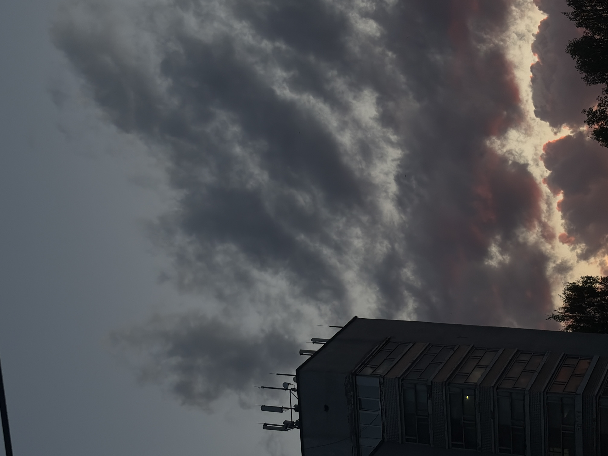 The image captures a low-angle view of a building against a dramatic sky. The building, located in the city of Unknown, Unknown, appears to be a multi-story structure with rows of windows and communication equipment mounted on its roof. The sky is filled with dark, heavy clouds, some tinged with red from the setting sun. A few trees are visible to the right of the building.