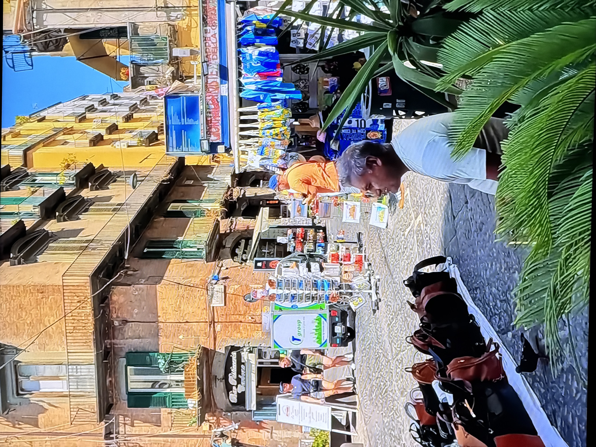 The image depicts a street scene in Chirchik, Uzbekistan, with several buildings featuring multiple balconies and awnings. The street is paved with cobblestones, and there are various vendors and shops. In the foreground, a man in a white t-shirt is seated, looking to his left. In front of him, a white tarp is spread out, displaying numerous backpacks in various colors, including red and black. A black bird is walking on the tarp.
To the left of the man, a small, white truck with a green logo that says "T group" is parked. Next to the truck, two women, one in a blue t-shirt and shorts, and the other in a black t-shirt and shorts, are standing and looking towards the truck. A white easel with a sign is placed to their left.
Further back, an orange-shirted man is standing near a stall displaying various colorful bags and clothing items hanging on a rack. Behind him, a shop with a sign reading "Bar Pasticceria" is visible. The buildings are multi-story, with brick facades and windows with green shutters on some of them. The sky is clear and blue.