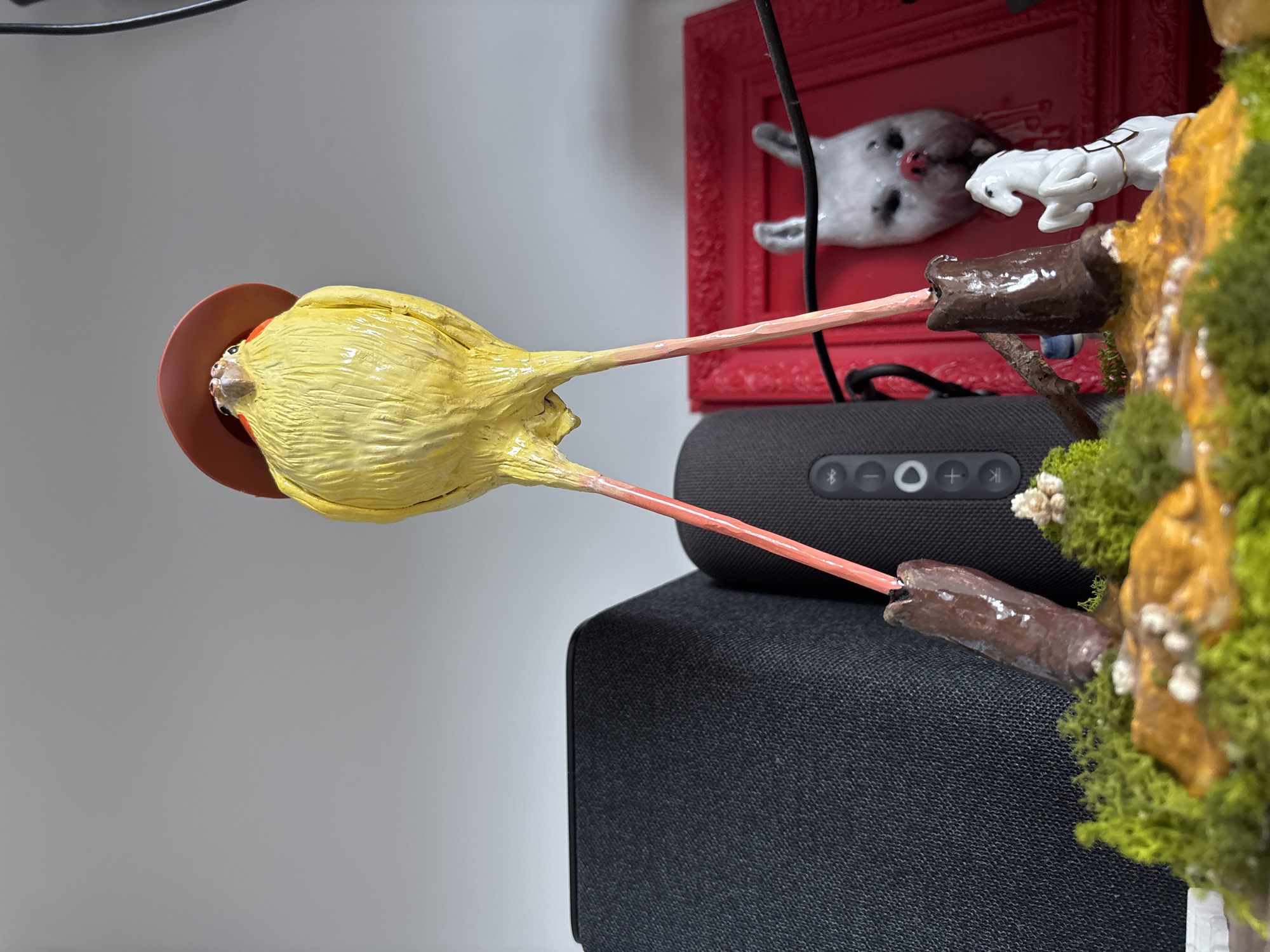 A close-up shows a whimsical, handcrafted sculpture of a yellow bird wearing a red hat. The bird appears to be balancing on two stilts, each resting on different surfaces. The left stilt is positioned on a black, rectangular speaker, while the right stilt sits atop a small diorama of miniature figures and moss. A red picture frame with a depiction of a grey and white animal is in the background. The scene is located indoors, against a light grey wall. The location in the city of Krasnoyarsk, Russia cannot be determined from the image.