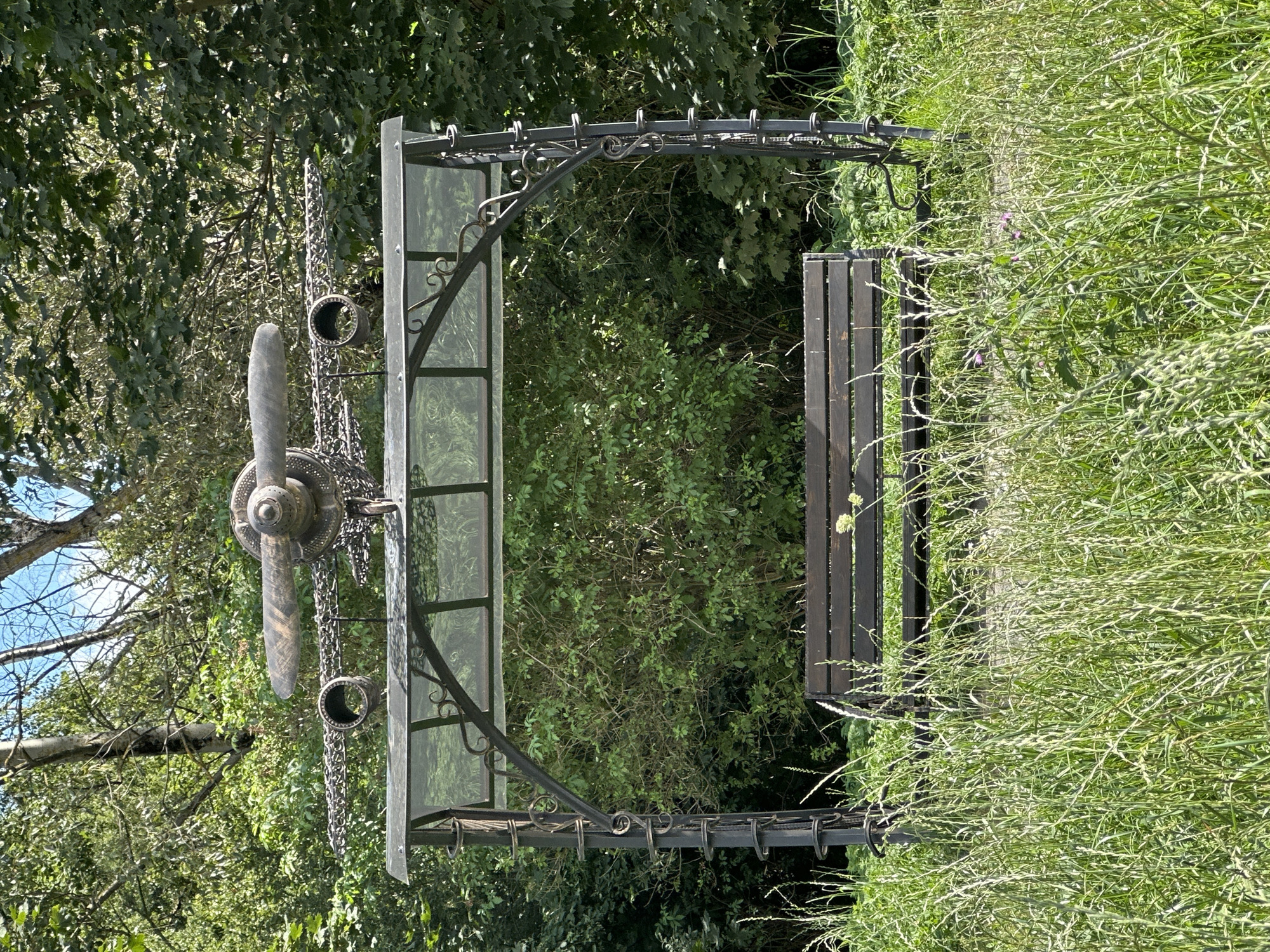 The image shows a metal bench with a canopy overhead, decorated with a propeller and wing-like structures resembling an airplane. The bench is situated in a park-like setting with abundant green foliage and tall grass in the foreground. The sky is visible through the trees in the background. There are no people or animals present in the scene. This artwork is located in Minsk, Belarus.