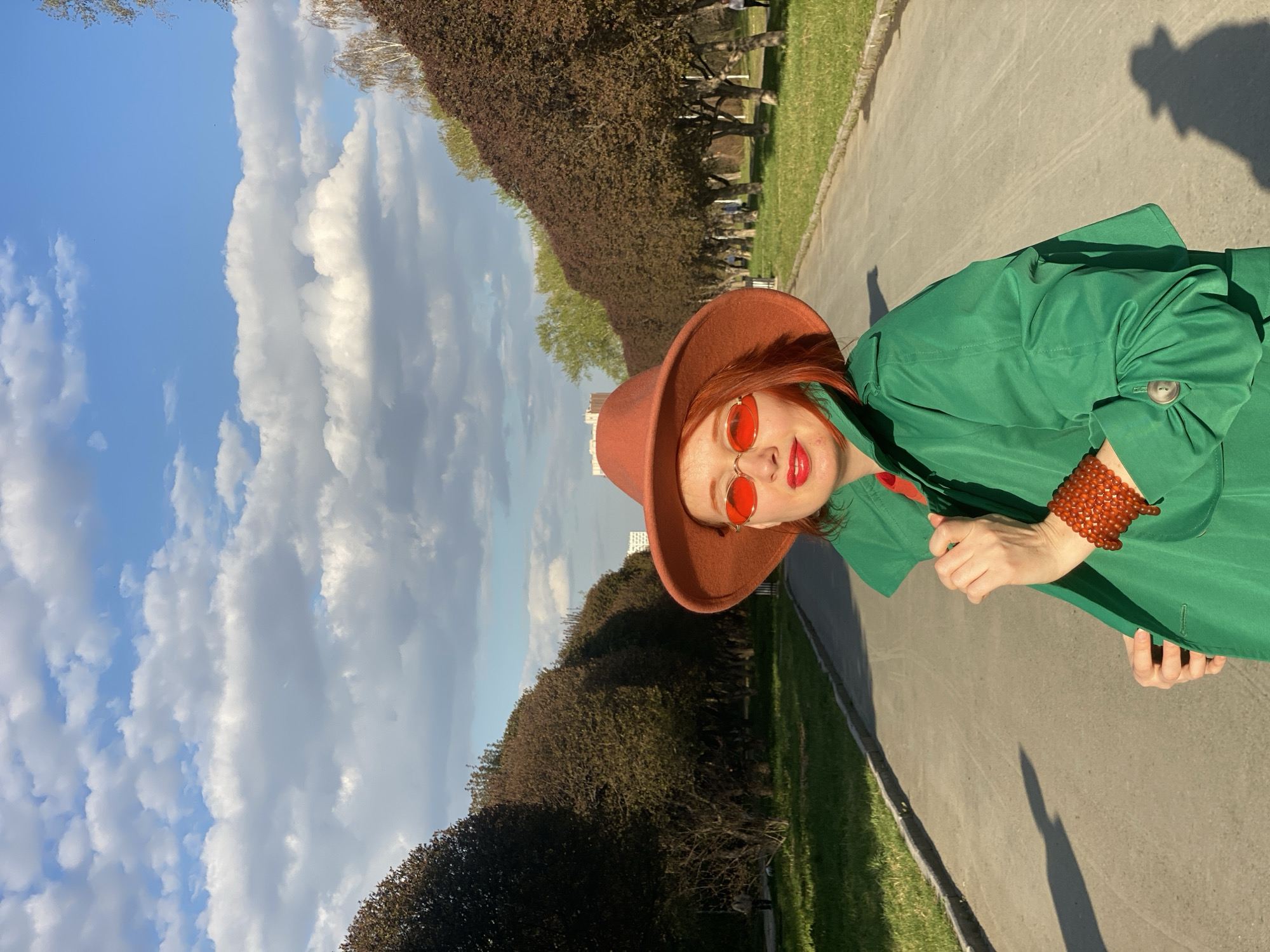 The photograph depicts a person, presumably a woman, outdoors on a sunny day. She is dressed in a green jacket and a brown hat, and she wears red-tinted sunglasses and a beaded bracelet. She has red hair and red lipstick. The person is positioned on a paved pathway. On either side of the pathway are rows of trees or bushes, forming a sort of avenue. In the background, a bright blue sky with fluffy white clouds is visible. The shadow of the person and her hat is cast on the pavement in front of her. The exact location is not directly discernible from the image alone. However, given the request for location to be identified in the city of Yekaterinburg, Russia, this photograph may have been taken in a park or garden in the city of Yekaterinburg, Russia.