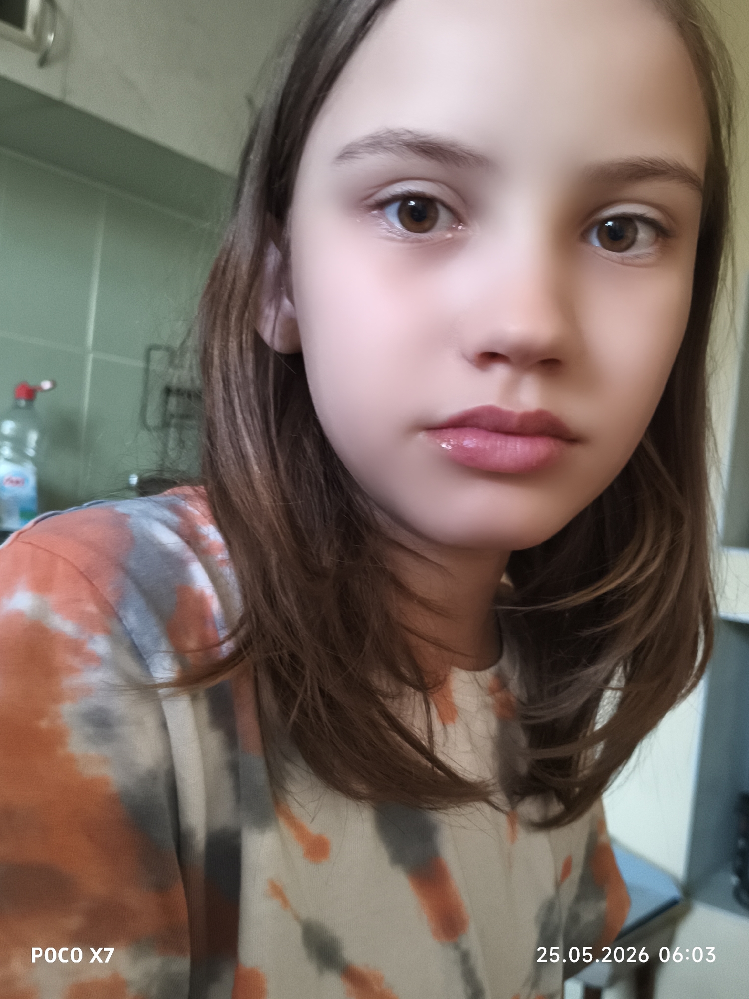 A young girl with brown hair and brown eyes is centered in the frame, wearing a tie-dye shirt with orange, gray, and white colors. Her lips appear to have a glossy finish. In the background, there is a light green tiled wall with a visible water bottle and a metal rack. A dark object, possibly a piece of furniture, is partially visible on the right side of the image. "POCO X7" and "25.05.2026 06:03" are printed at the bottom of the image. The location is Unknown, Unknown.