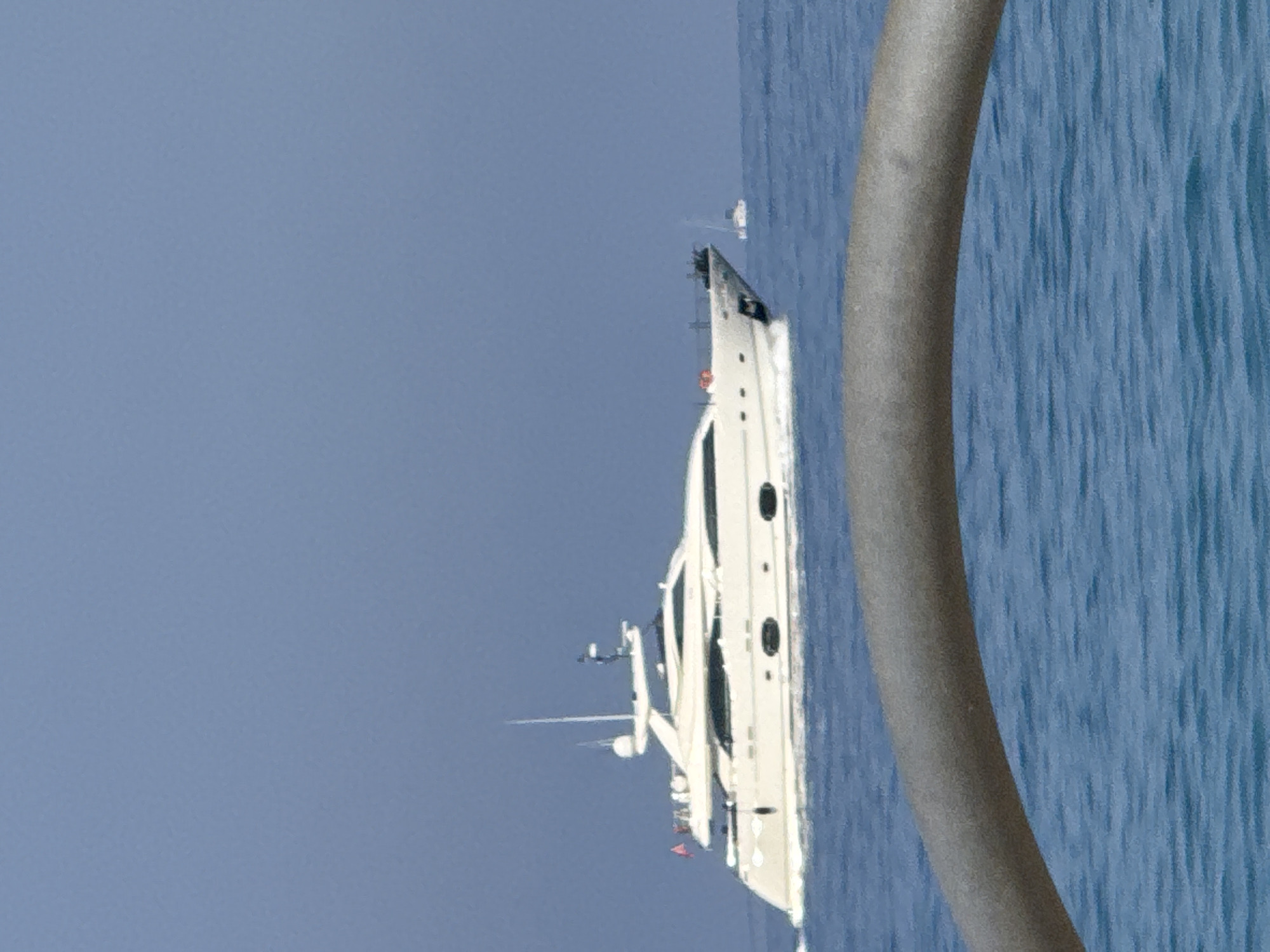 A wide-angle, eye-level shot captures a large, white yacht sailing on the blue sea under a clear, pale blue sky. The yacht is the primary subject, positioned in the middle ground and moving from left to right. It features multiple decks, windows, and a tall mast. A smaller white boat is visible in the distance on the right. In the foreground, a curved, metallic railing or structure partially obstructs the view of the water, which appears choppy. The image does not provide any specific visual cues to pinpoint a location within Fethiye, Turkey.