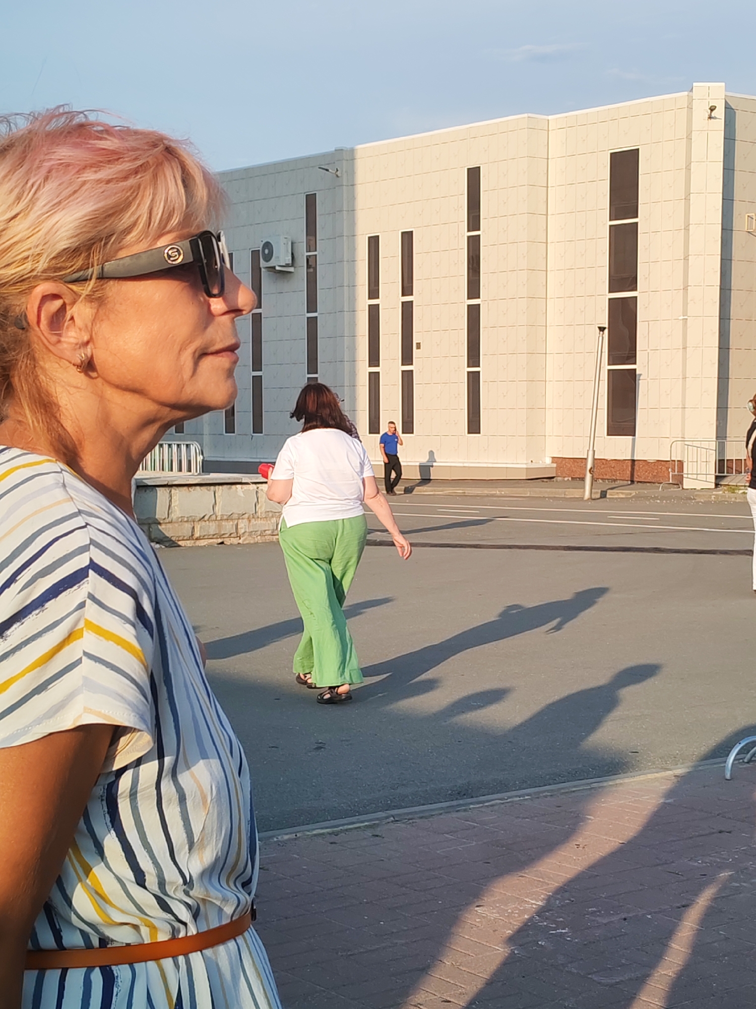 In an outdoor, urban setting, a woman with light-colored hair with pink highlights, wearing sunglasses and a horizontally striped dress, is in the foreground, her profile facing right. In the middle ground, a woman in a white t-shirt and green pants walks away from the camera. Behind her, a man in a blue shirt and black pants stands near a building, talking on his phone. The background is dominated by a modern, beige building with dark windows and what appears to be an air conditioning unit. The scene is bathed in sunlight, casting long shadows on the asphalt and brick walkway.