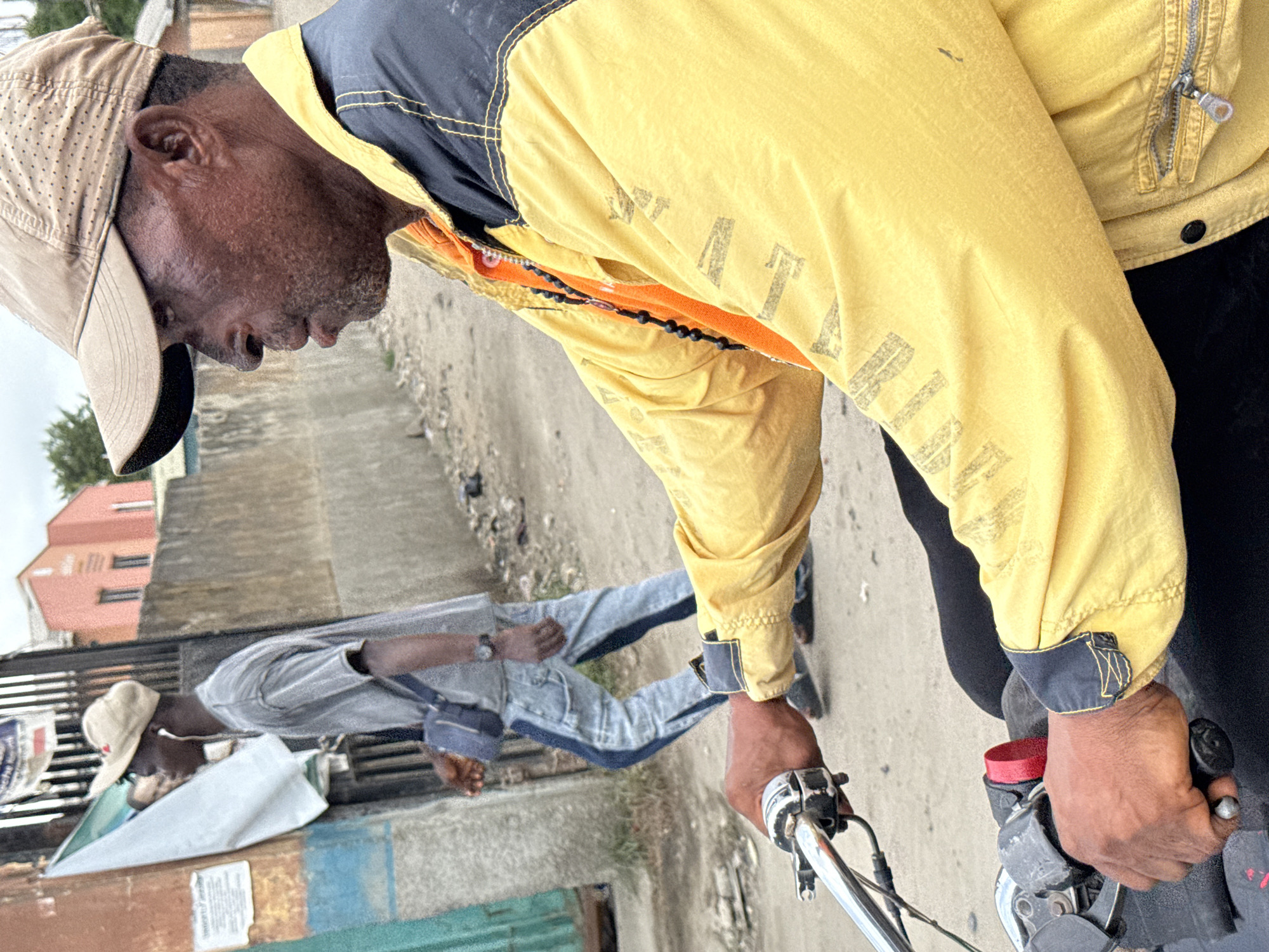 A medium shot depicts an adult Black man on a motorcycle in Lekki, Nigeria. He wears a tan baseball cap and a yellow jacket with black accents. His hands grip the motorcycle's handlebars. Another man, also Black, wearing a light gray shirt and matching pants with dark trim and a tan cap, stands in the background. A blue bag hangs across his shoulder. The setting appears to be a street, with buildings visible in the background. The scene is slightly blurry.