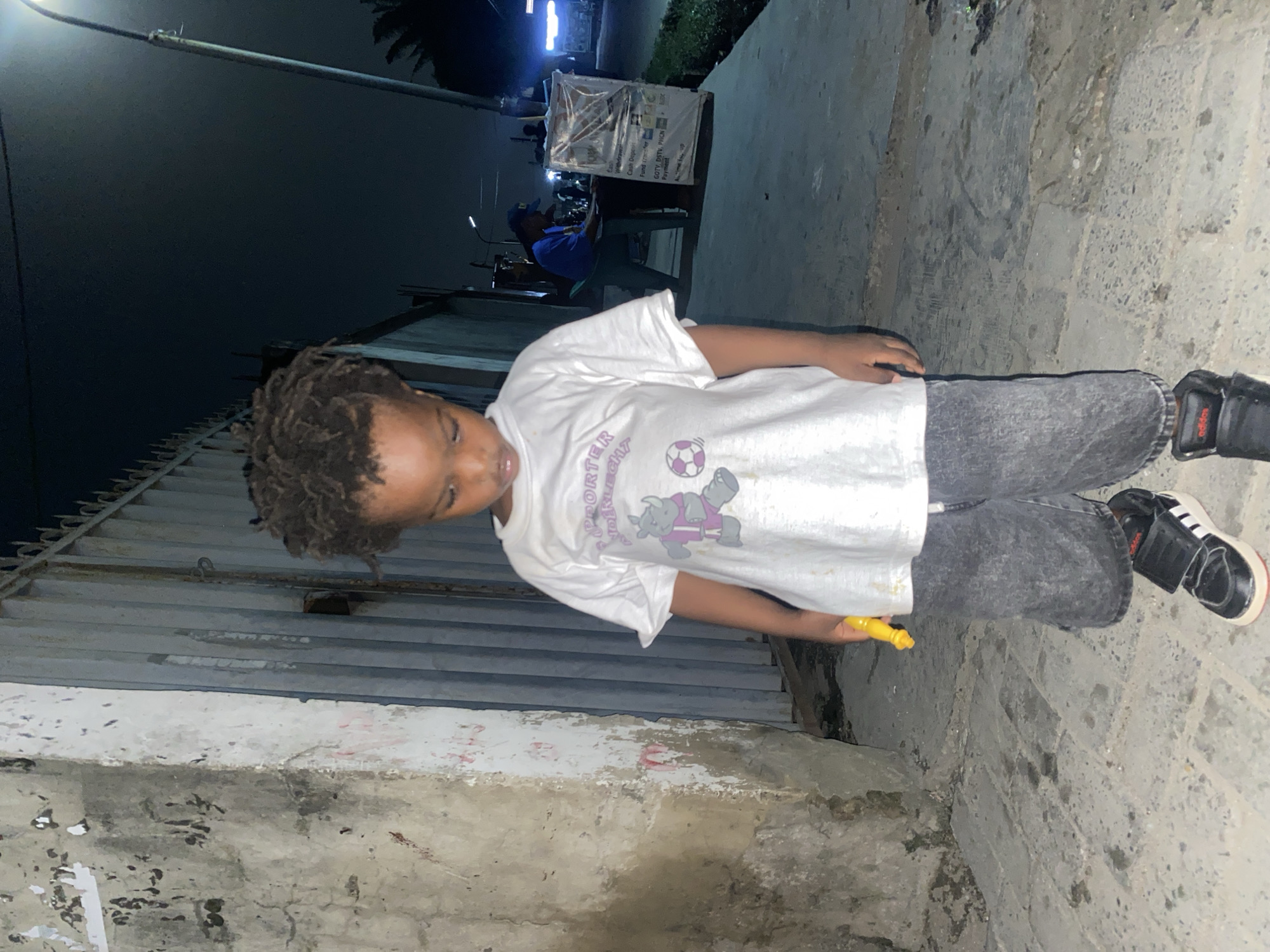 A young child stands outdoors on a paved surface that may be a sidewalk or street in Ado, Nigeria. The child wears a white t-shirt with a cartoon graphic, gray jeans, and black and white sneakers. They hold a yellow object, possibly a toy, in their right hand. The background includes buildings of rough concrete and corrugated metal sheets. The scene is illuminated by a street light, suggesting it is night. A distant, brightly lit booth or kiosk is visible in the background. A person is seated near the booth.