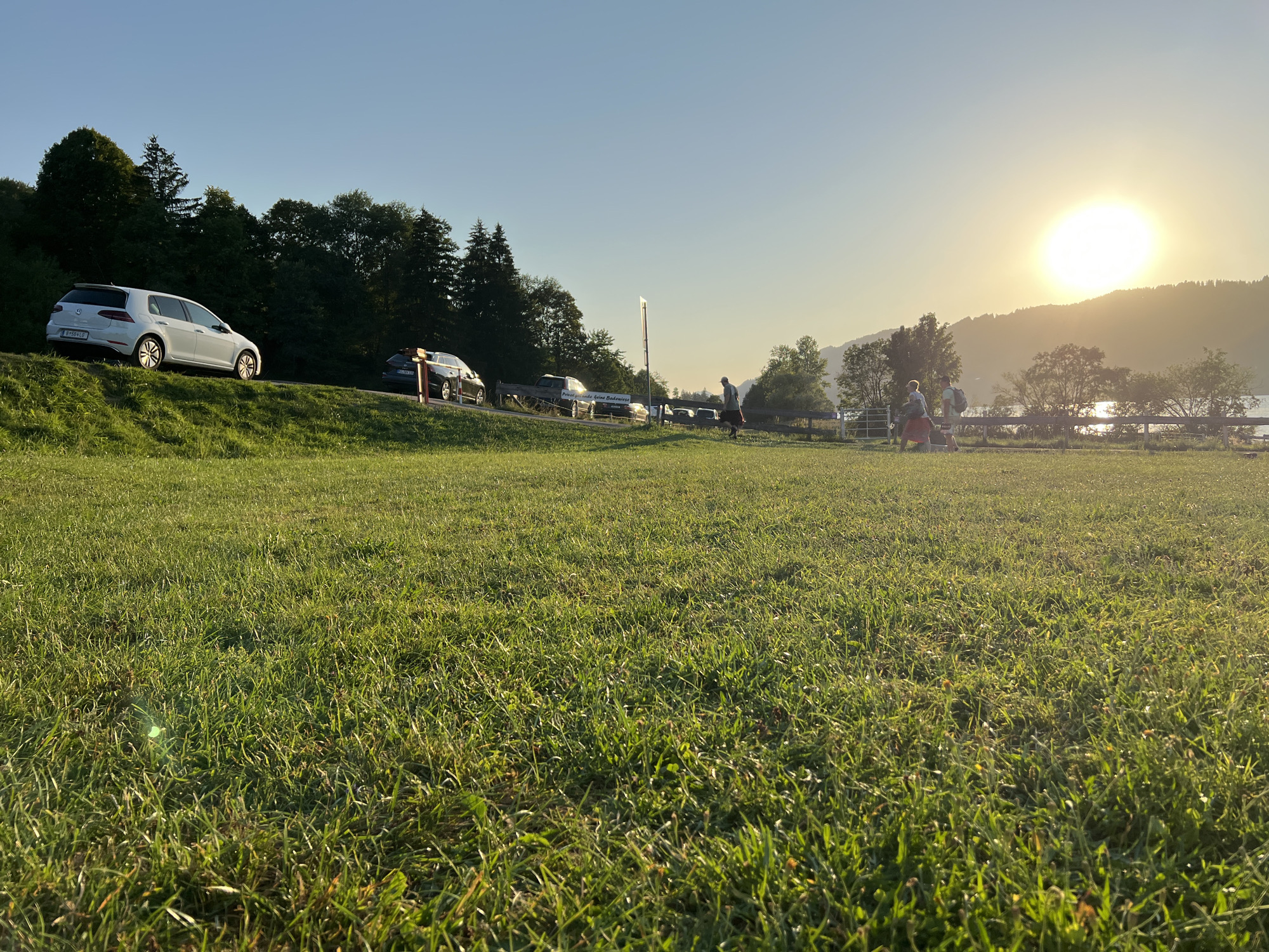 The image depicts a grassy field with several cars parked along a road in the background. A white car is parked on the left side of the road, facing away from the viewer. Further down the road, there are several other cars, including a dark-colored station wagon.
In the middle ground, a group of people are walking along a fence. The sun is setting in the background, casting a warm glow over the scene. The sky is clear and blue, with the sun appearing as a bright orb. The location is Immenstadt im Allgäu, Germany, indicated by the presence of a sign in German that reads "Private, keine Badewiese" which translates to "Private, no bathing meadow." The overall scene suggests a pleasant evening outing in a scenic, natural environment.